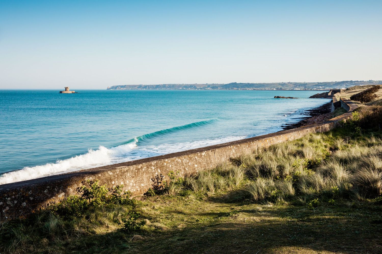 Panoramic view of a pristine coastline with turquoise waters and grassy cliffs on a clear summer day.