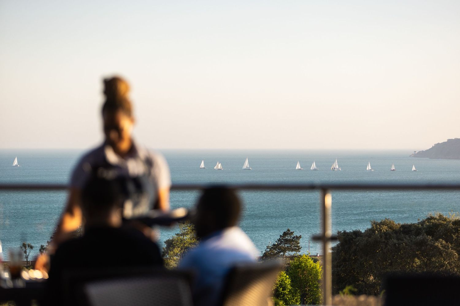 Panoramic ocean view with sailing boats and coastal homes in golden evening light.