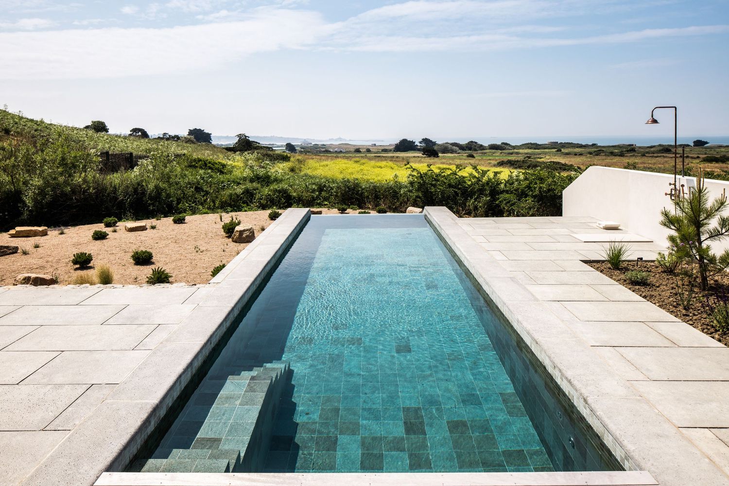 A modern infinity pool with turquoise water extends towards rolling hills and countryside views on a sunny day.