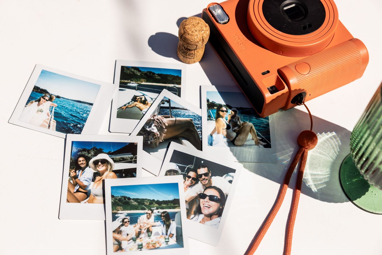 Orange instant camera with scattered vacation photos and cork on white surface.