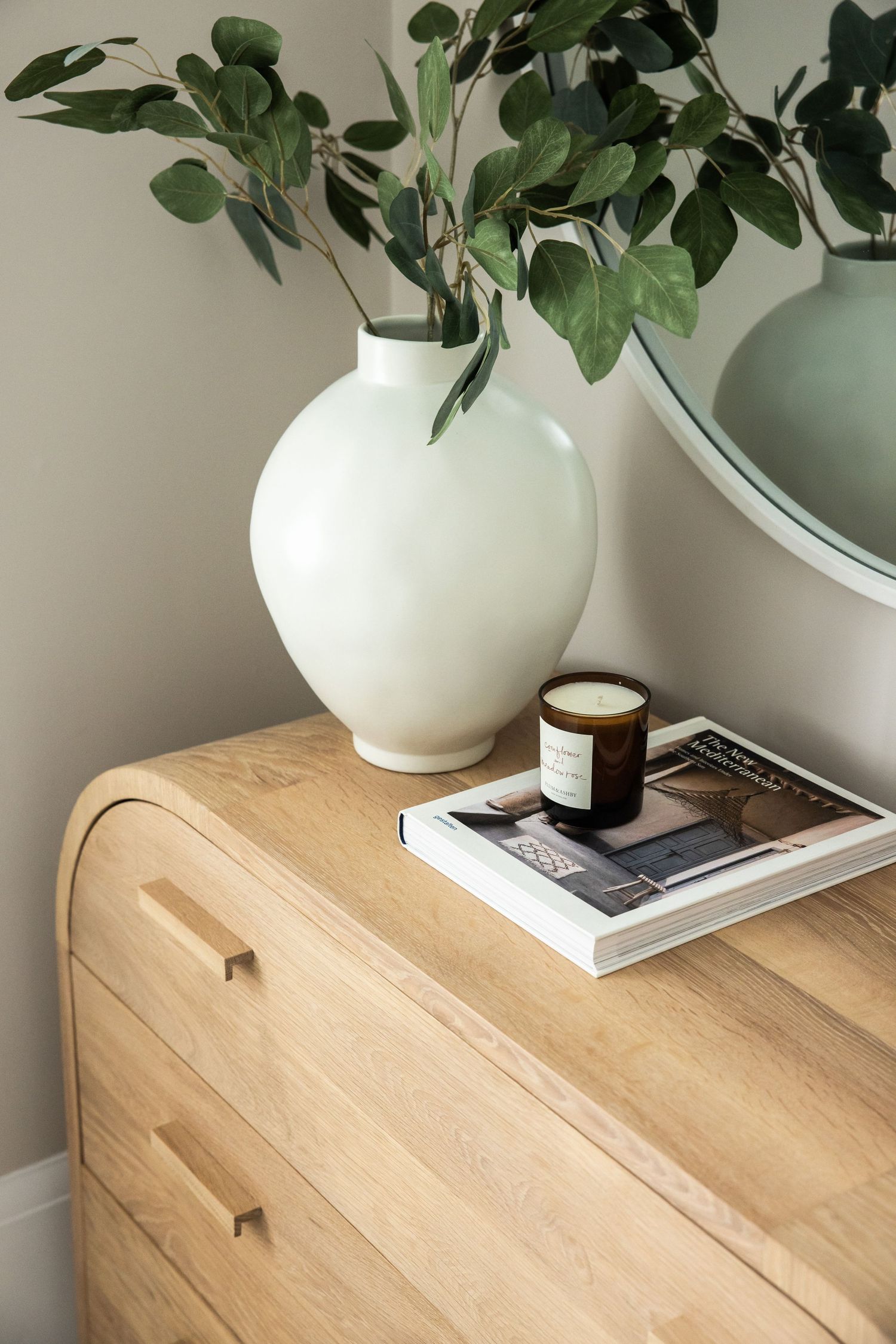 White ceramic vase with green leaves and book styling on wooden dresser surface.