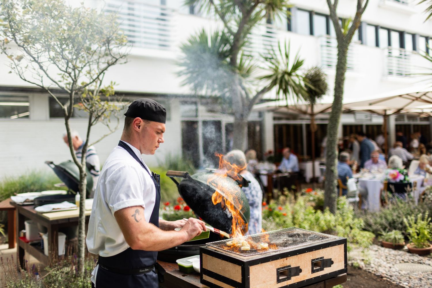 Chef preparing gourmet dishes at an outdoor poolside restaurant kitchen with palm trees in background.