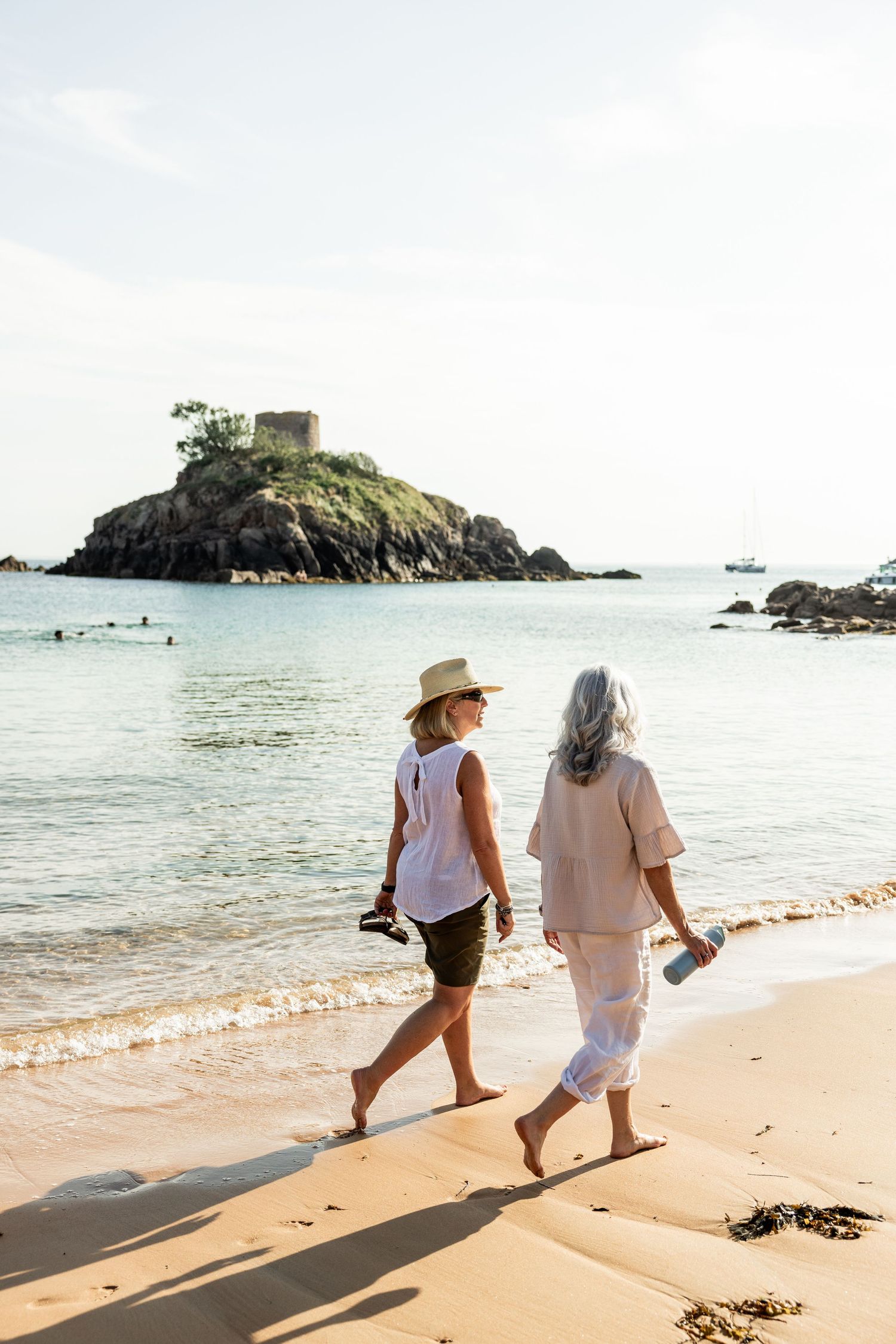 Two people walking along a sandy beach with a historic stone tower visible on the rocky coastline.