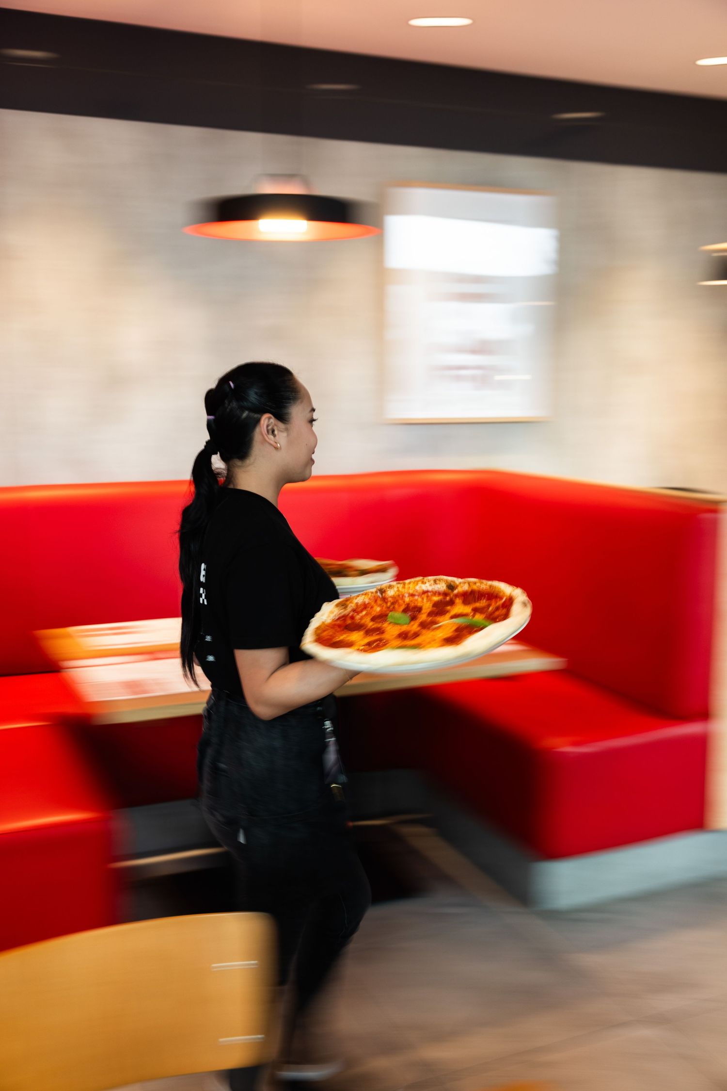 Server carrying a pizza plate through a restaurant with bright red booth seating.