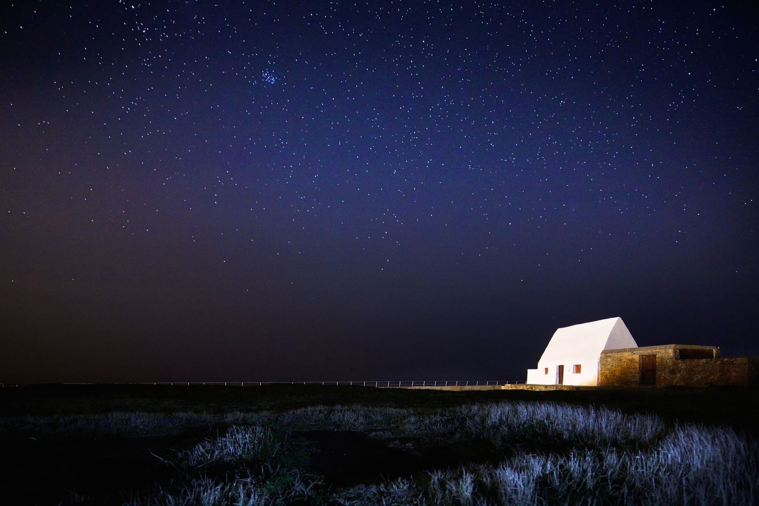 Starry night sky over a solitary white lighthouse perched on a rocky coastal cliff.