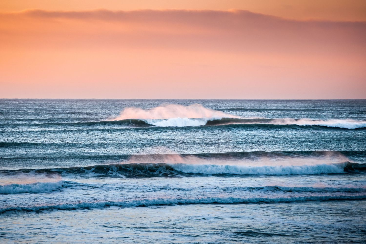 Ocean waves roll in during a pink sunset as spray catches the light against a pastel sky over the sea.