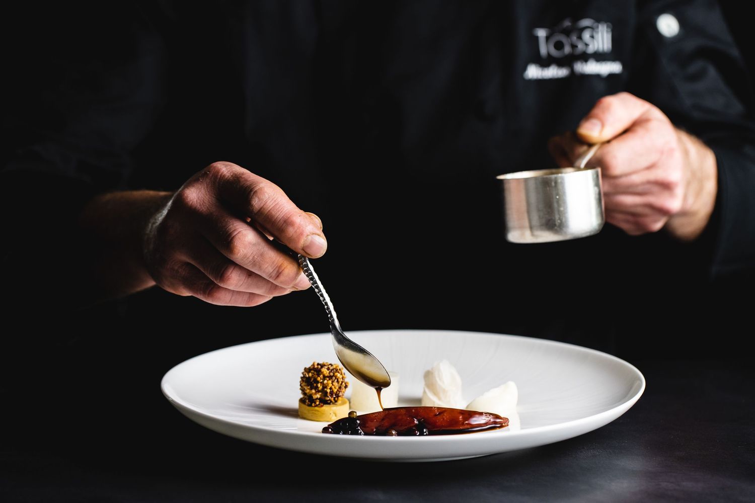 Chef adding finishing sauce to plated meat dish in fine dining setting.