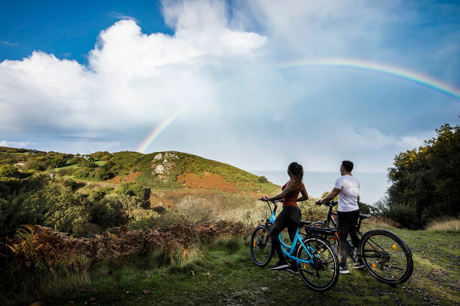Cyclists admiring rainbow over green hillside landscape.