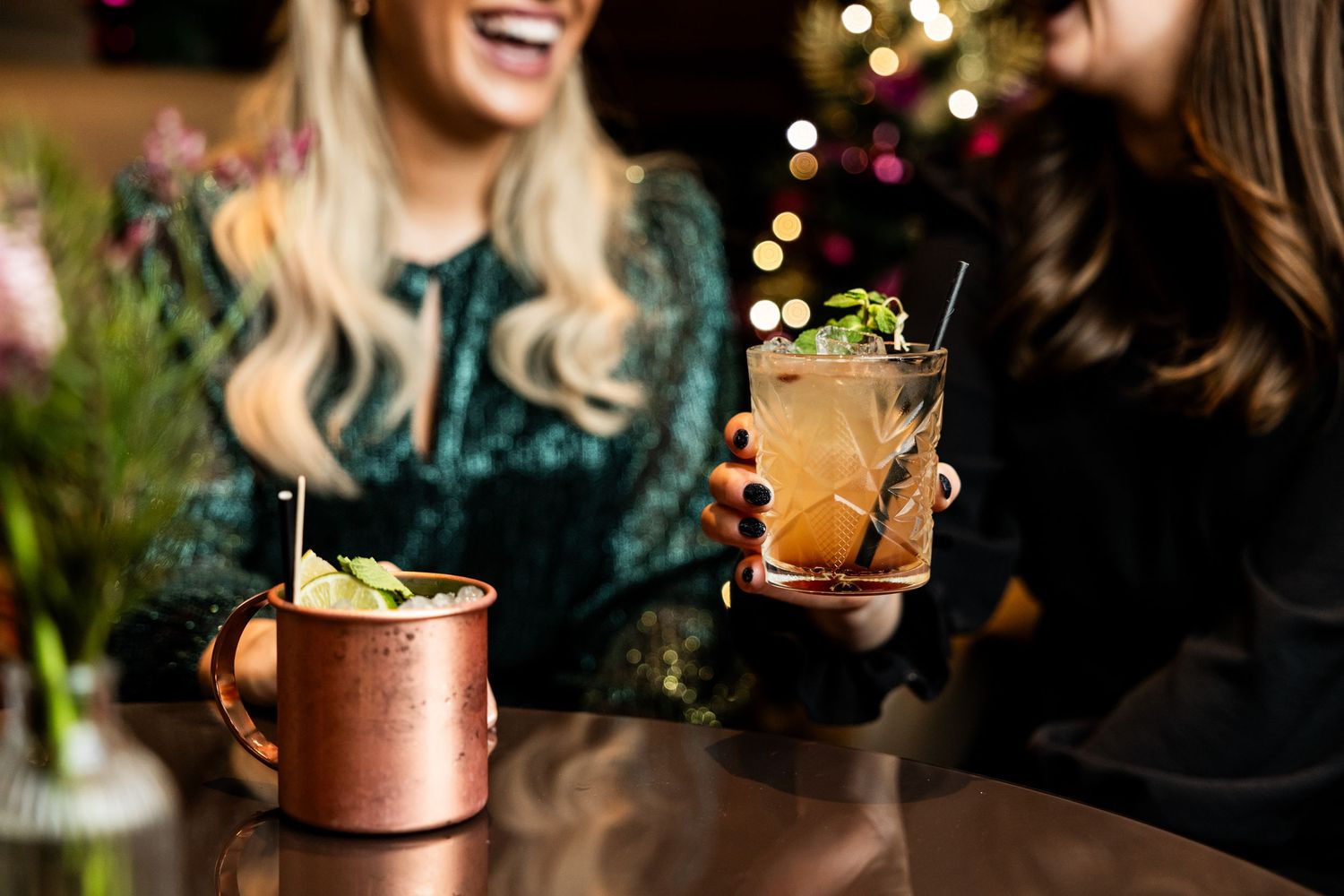 Two festive cocktails being served with garnishes and holiday lights in background.