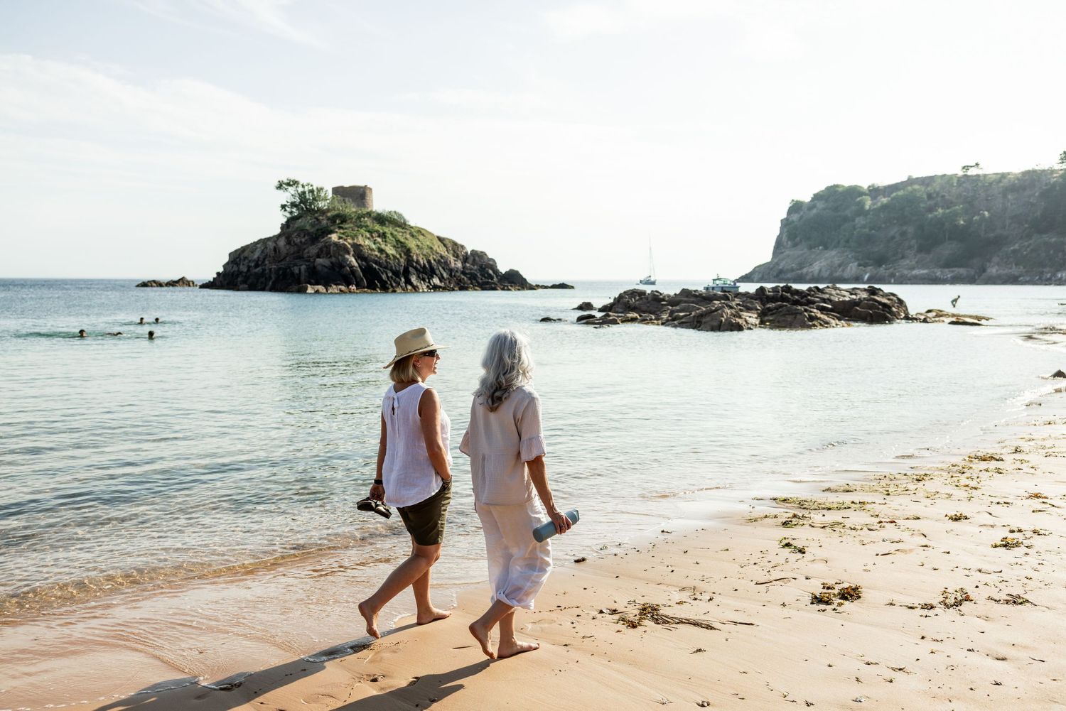 People walking along a scenic coastal beach path with rocky cliffs and blue ocean waters in the background.