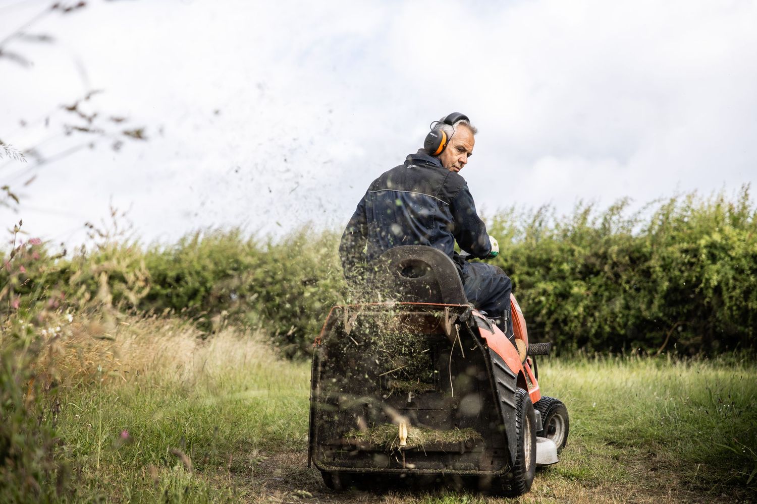 Riding lawn mower parked outside rural storage shed surrounded by tall grass.