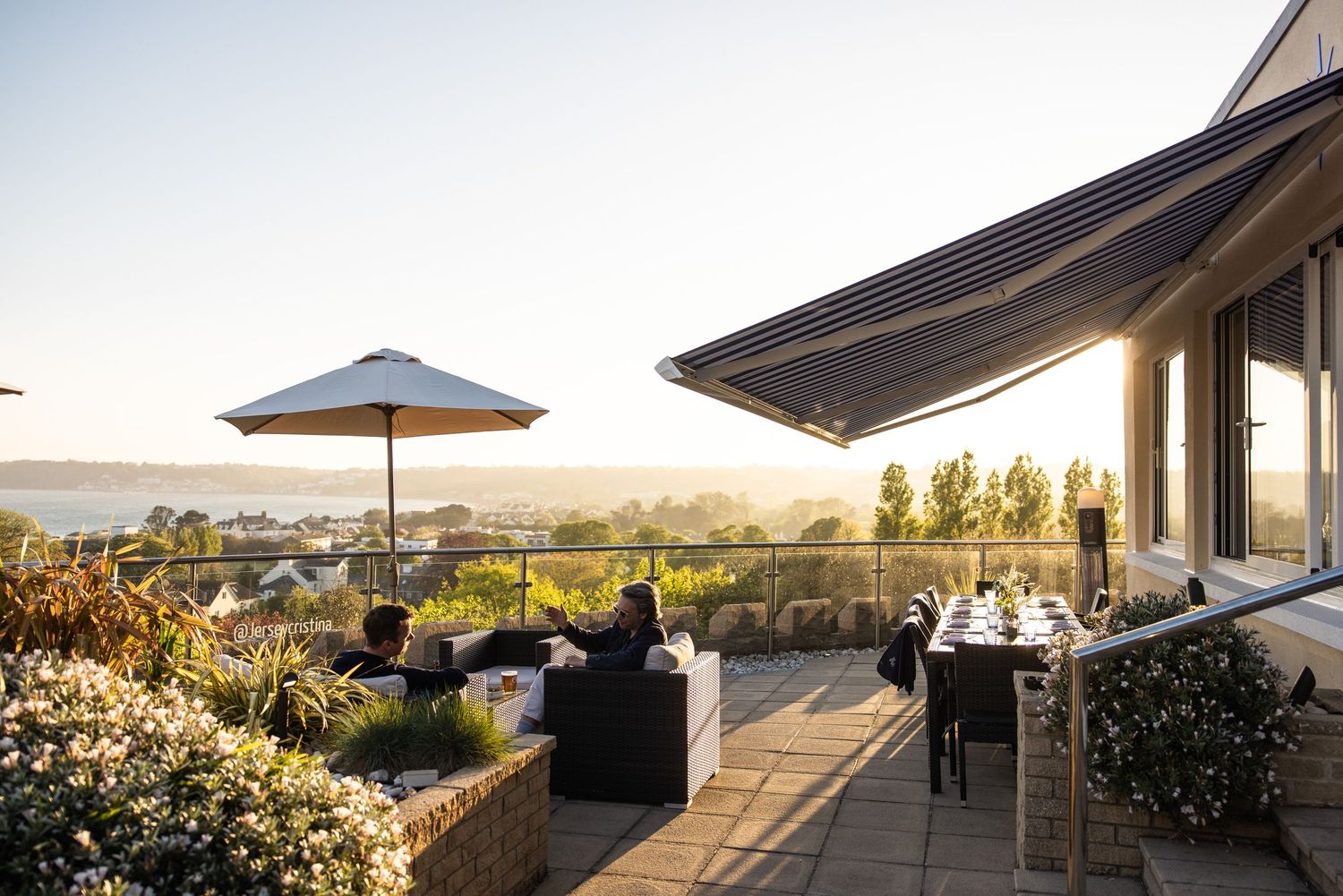 Outdoor terrace with umbrellas and seating overlooking a coastal landscape at sunset.