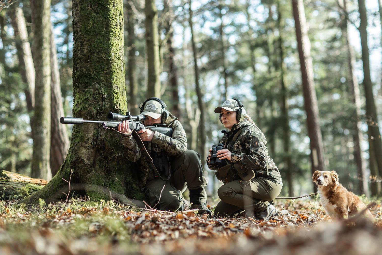 Two hunters in camouflage crouch by a tree in a forest, one aiming a rifle while a golden retriever sits nearby.