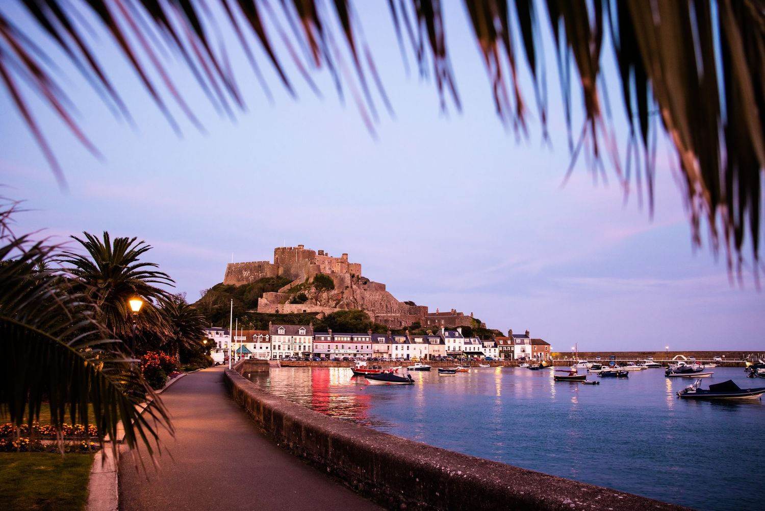 Panoramic view of a coastal town harbor with boats and castle at dusk.