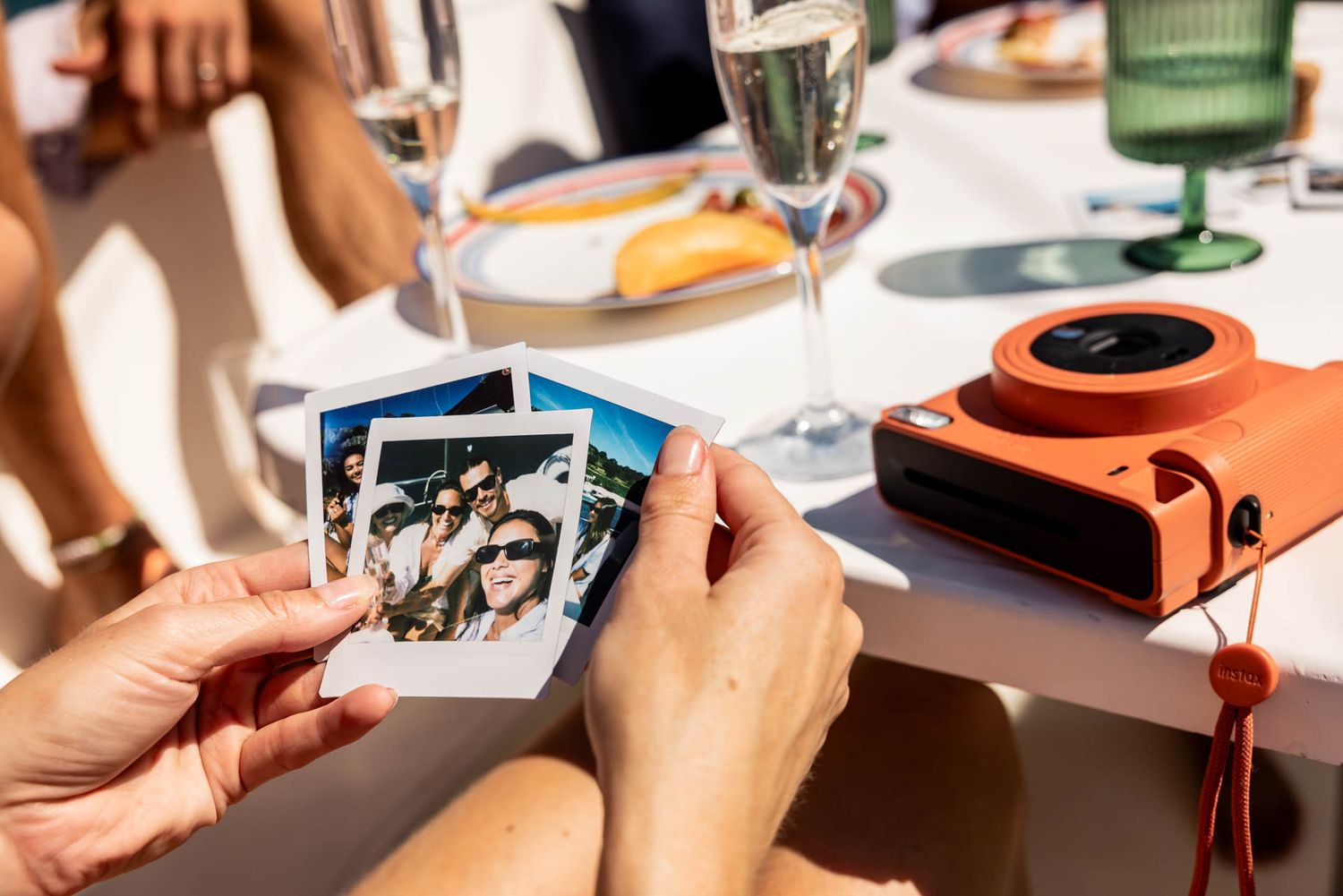 Hands holding instant photos at dining table with orange camera and wine glasses.
