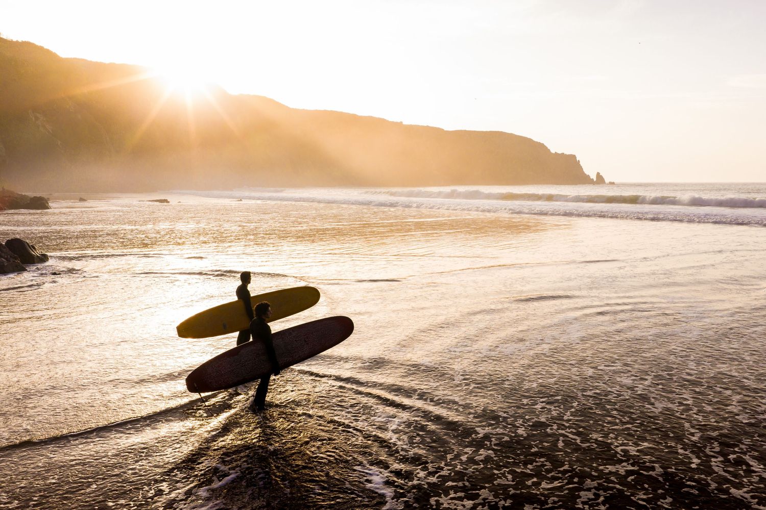Silhouette of surfer with boards walking along misty beach at sunset with coastal cliffs.