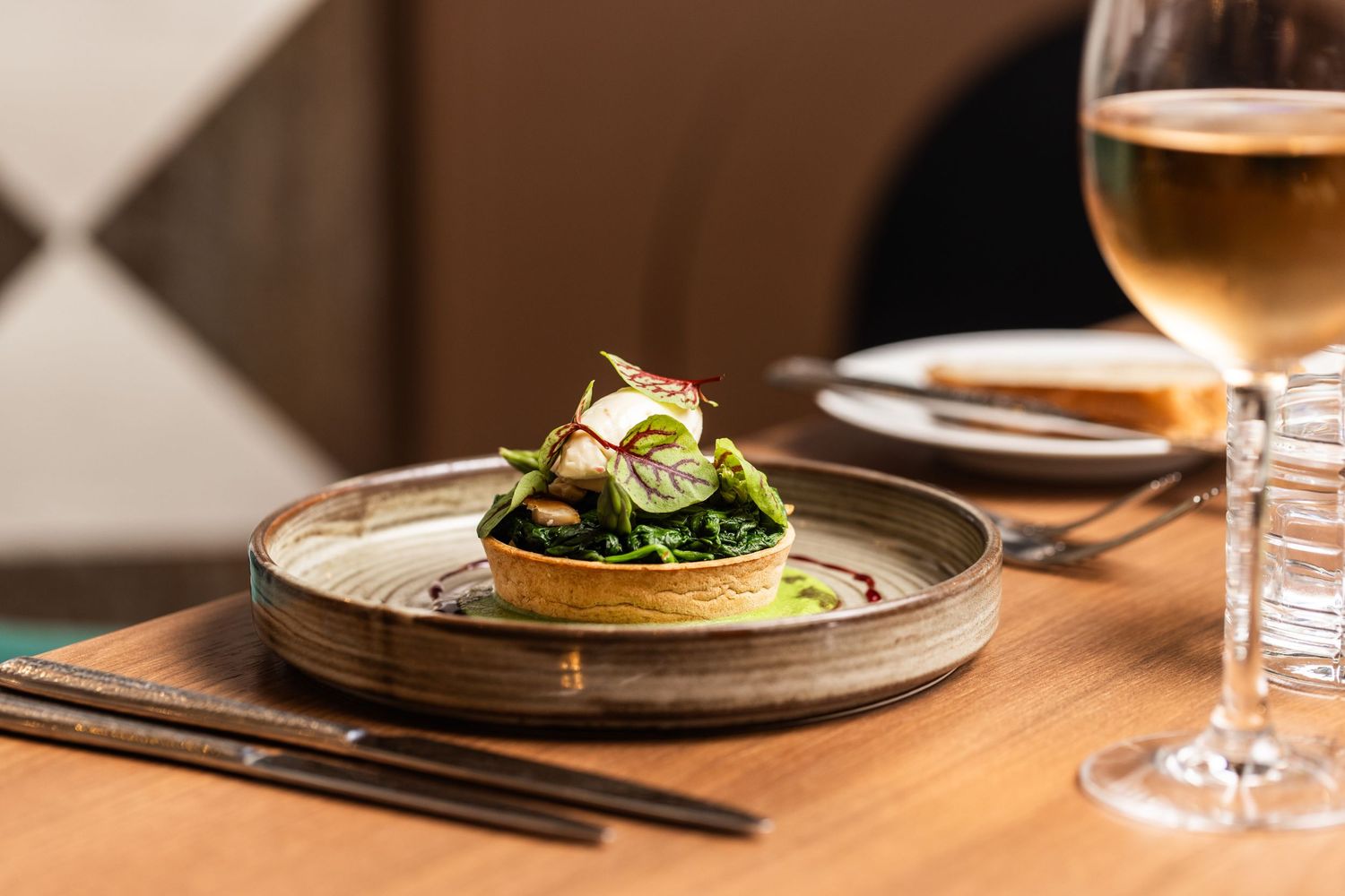 Elegant fine dining dish with sautéed greens on a ceramic plate, paired with a glass of white wine on a wooden table.