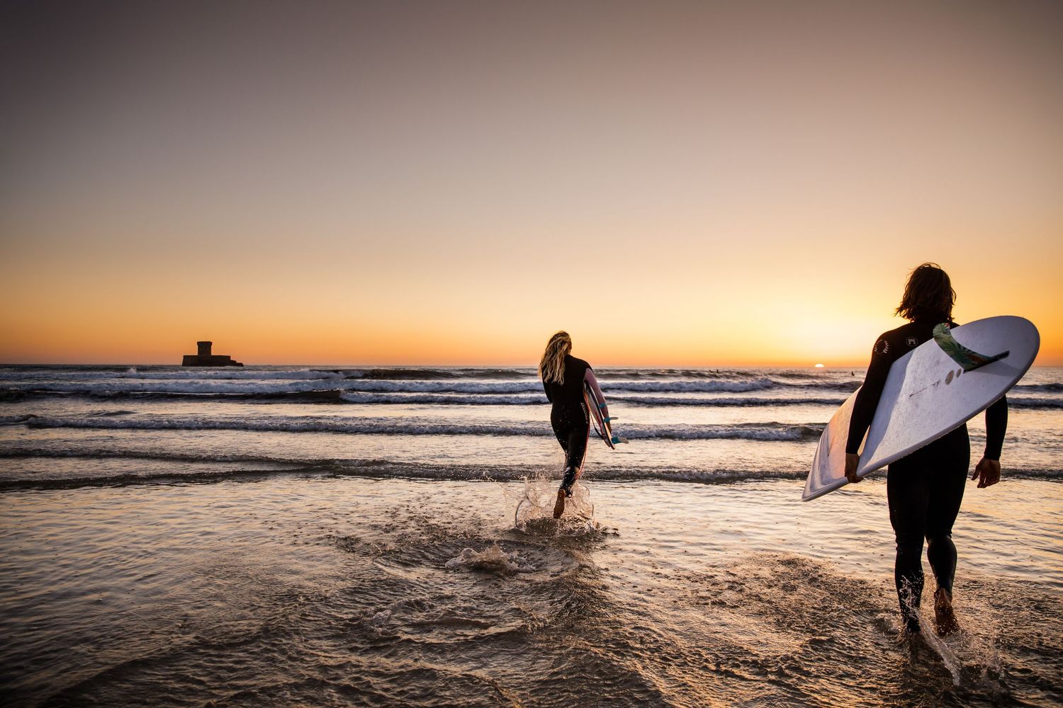 Silhouettes of surfers walking along a beach at sunset with surfboards and golden light reflecting on the ocean.