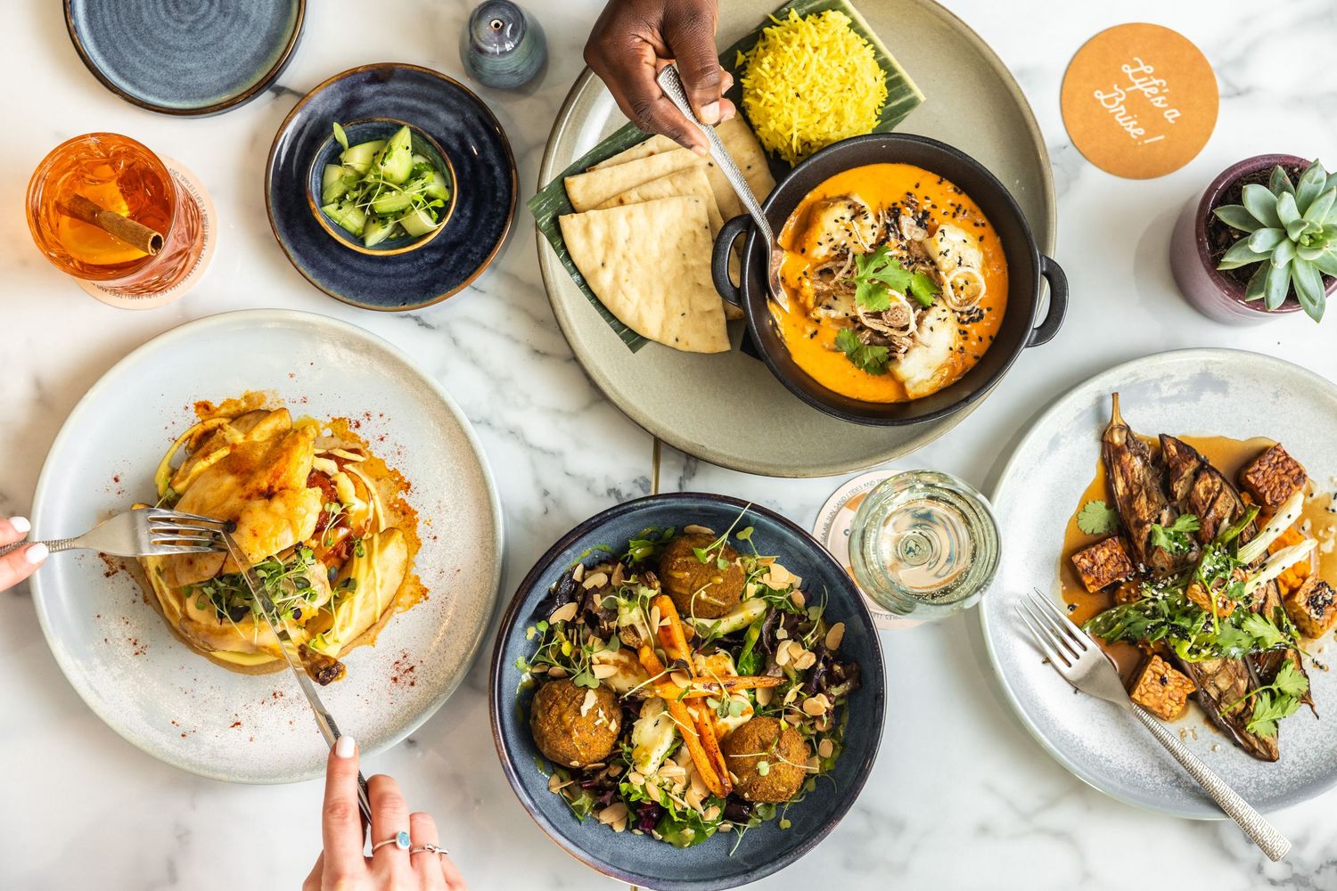 Overhead view of colorful restaurant dishes including curries, salads and appetizers on a light background.