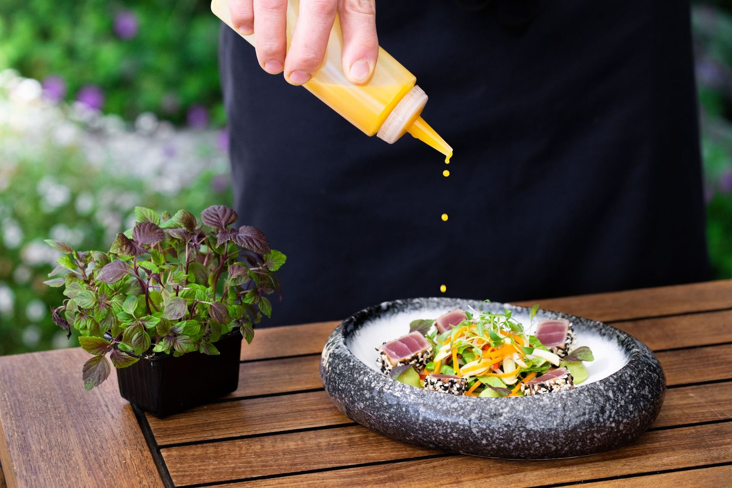 Orange sauce being drizzled over plated dish in outdoor garden setting.