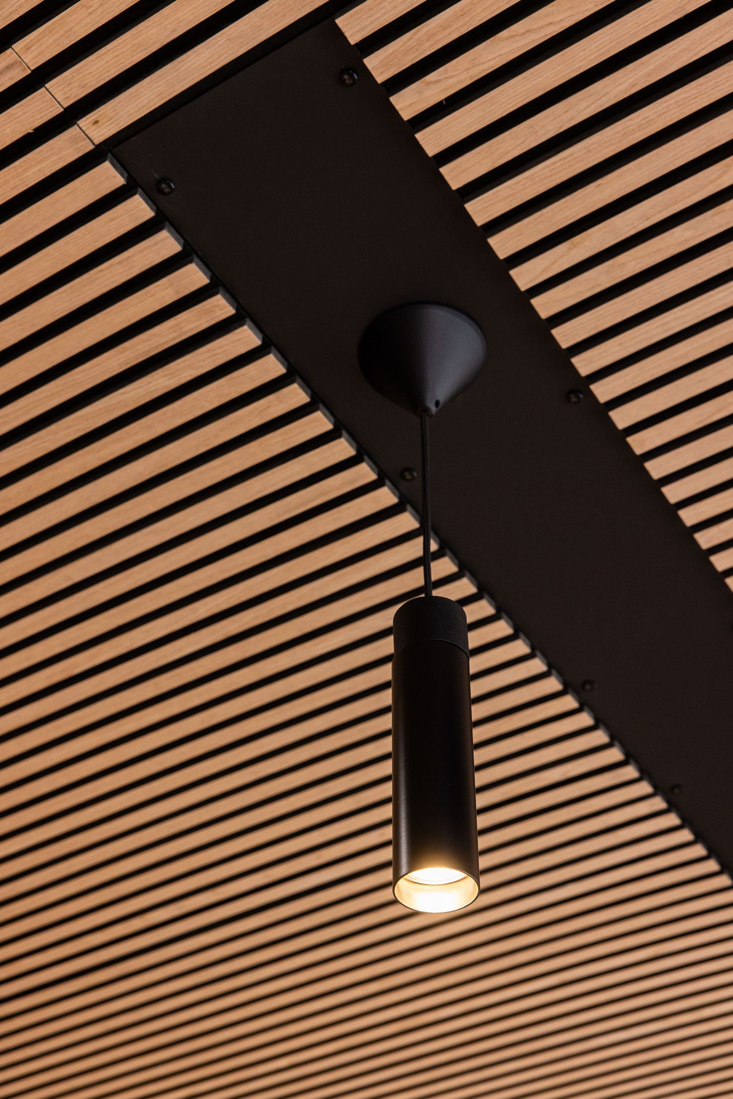 Cylindrical black pendant light hanging from wooden slatted ceiling with geometric pattern.