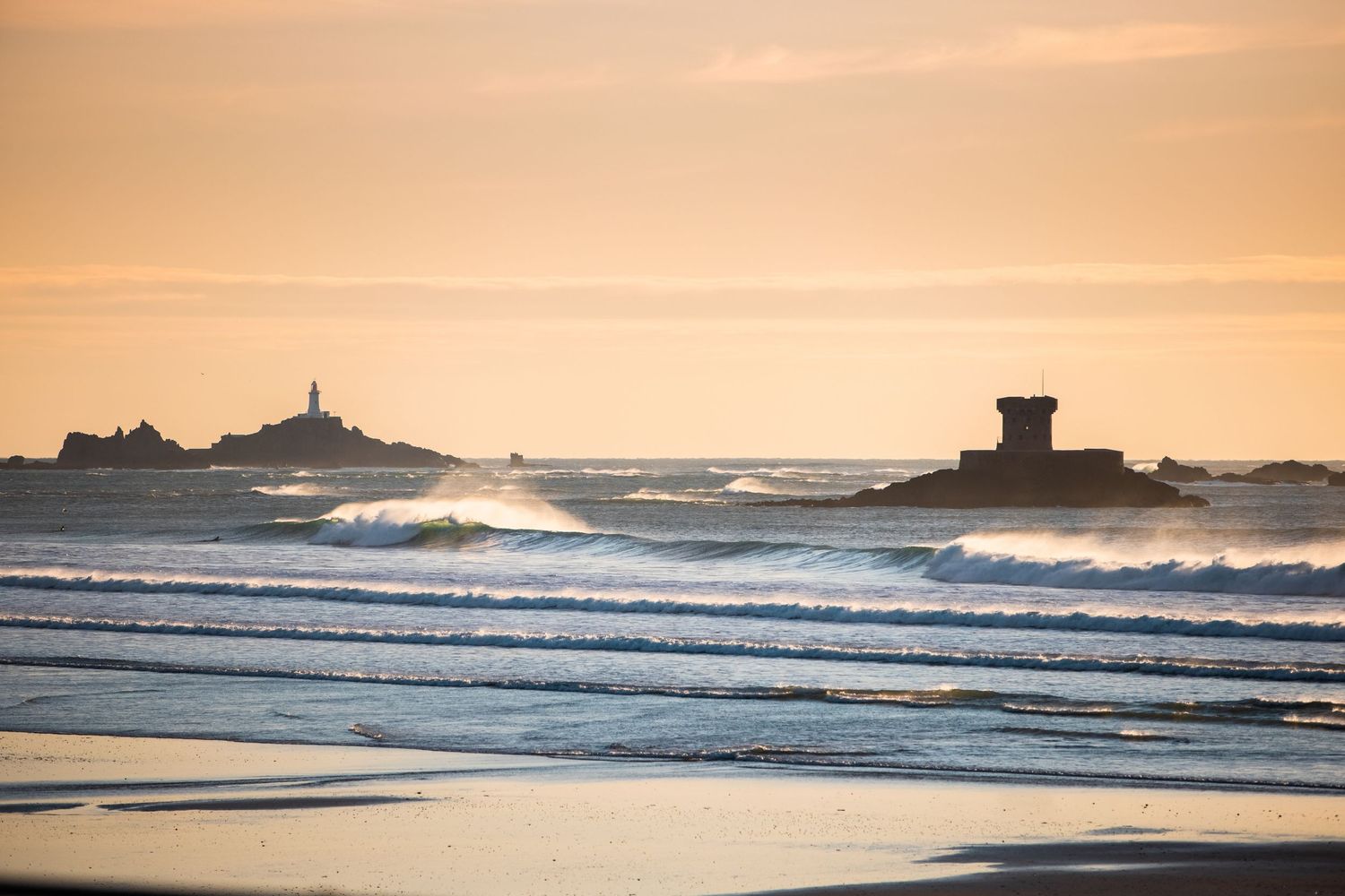 Golden sunset illuminates the ocean waves breaking near a distant coastal lighthouse.