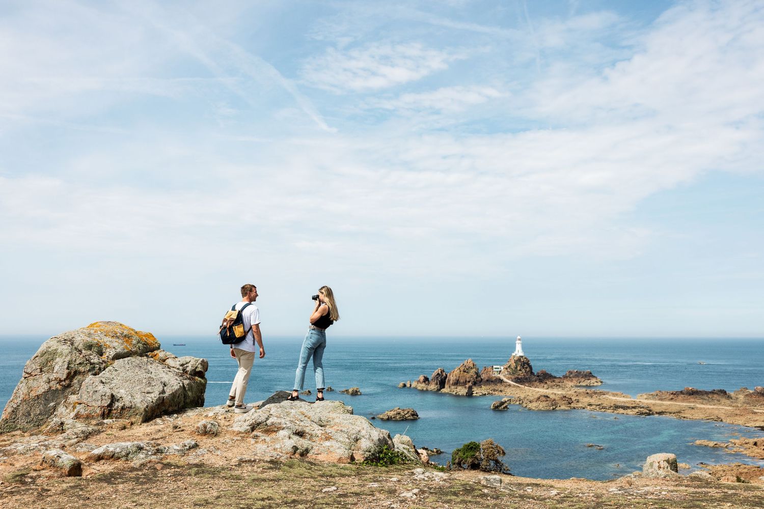 Rocky coastal landscape with sailboats visible in the distance under blue skies.
