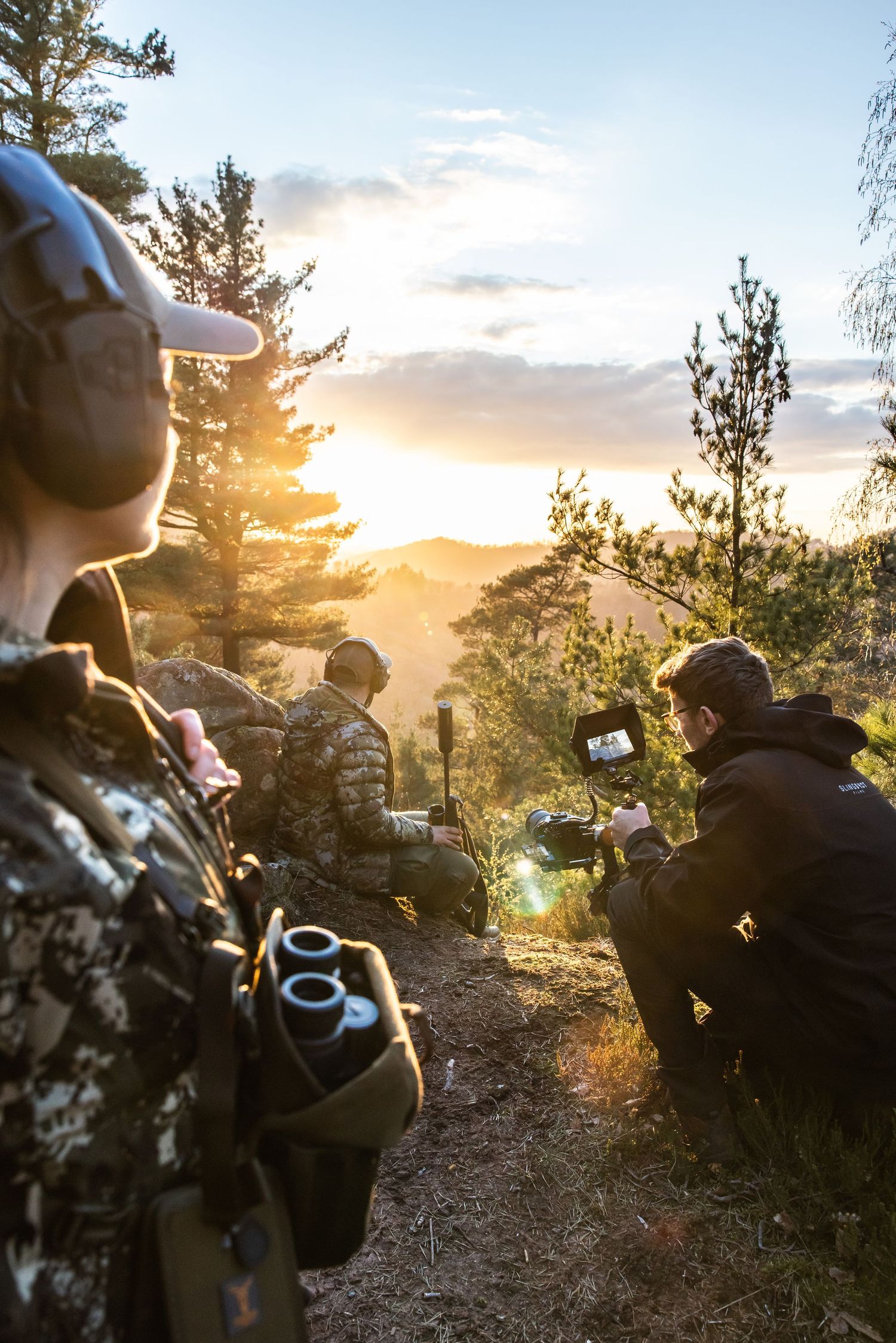 Hunters in camouflage gear film a scenic golden sunset over forested mountains using a camera stabilizer.
