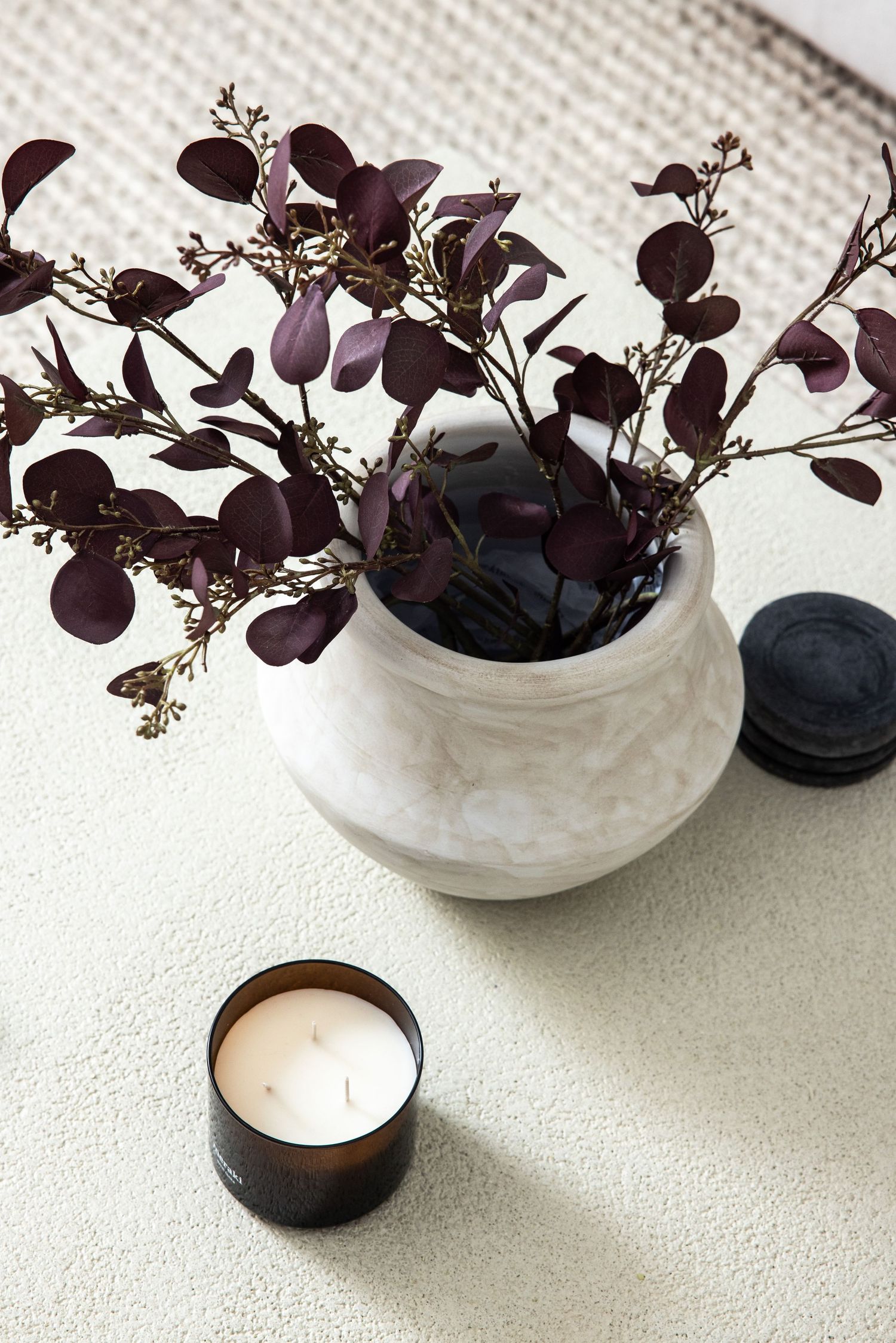Purple floral arrangement in white vase with small candles on white surface.