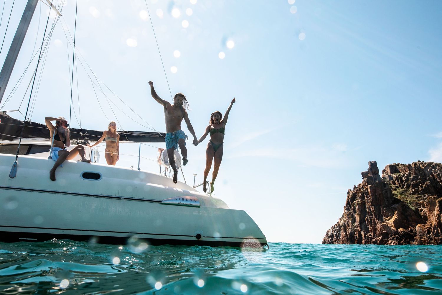 Silhouettes of people jumping from a catamaran yacht in crystal blue waters.