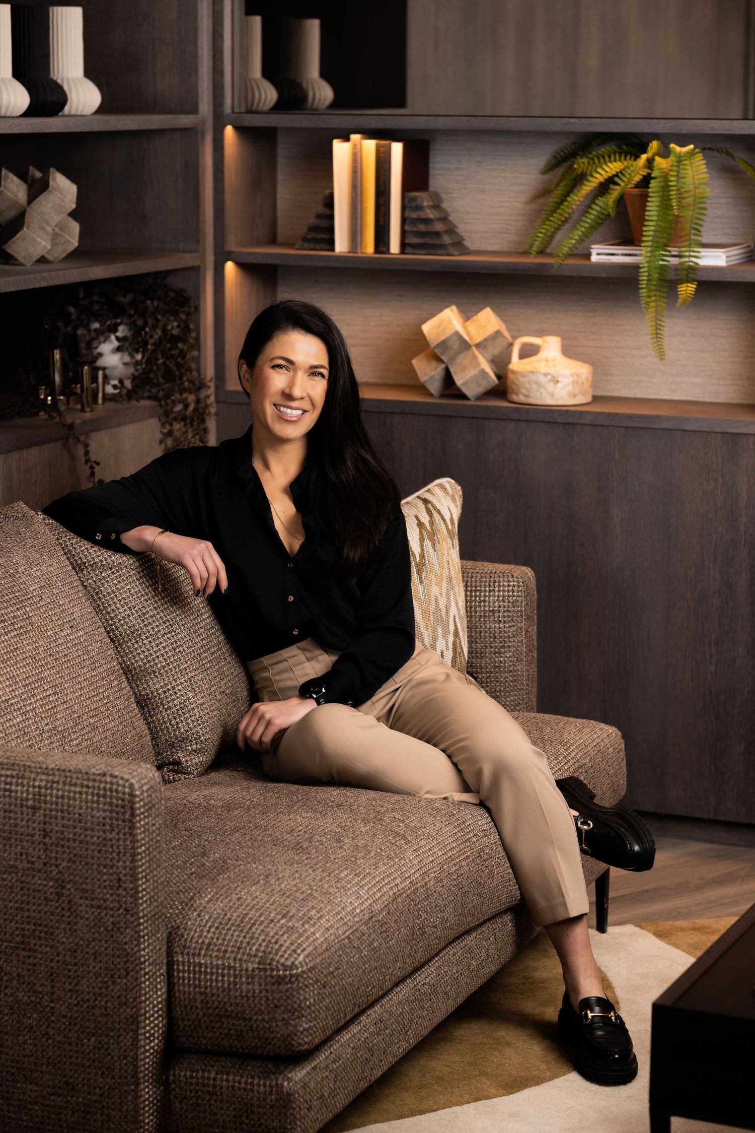 Professional in black shirt relaxes on gray couch with decorative shelving in background.