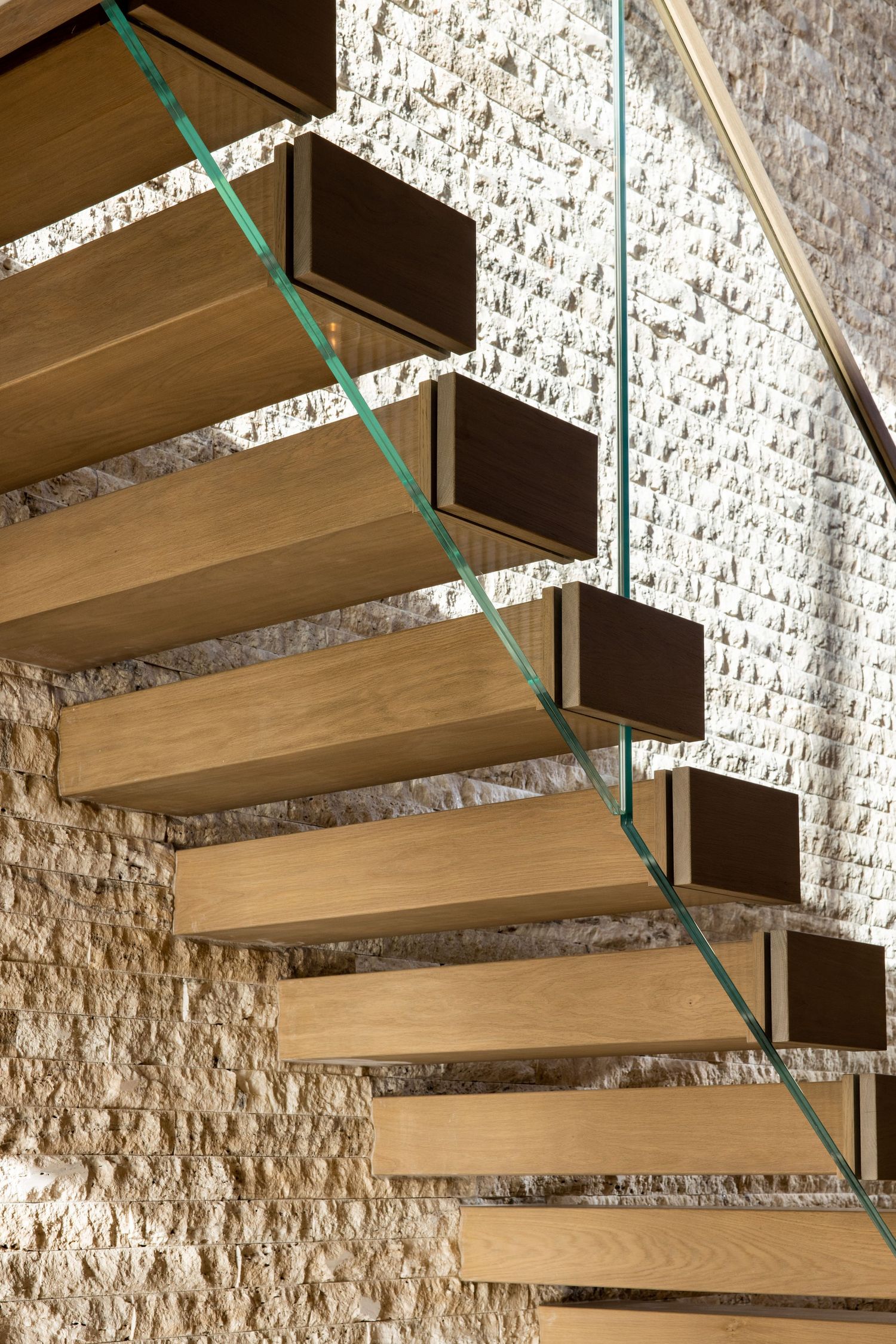 Floating wooden staircase with glass railing against textured limestone wall.