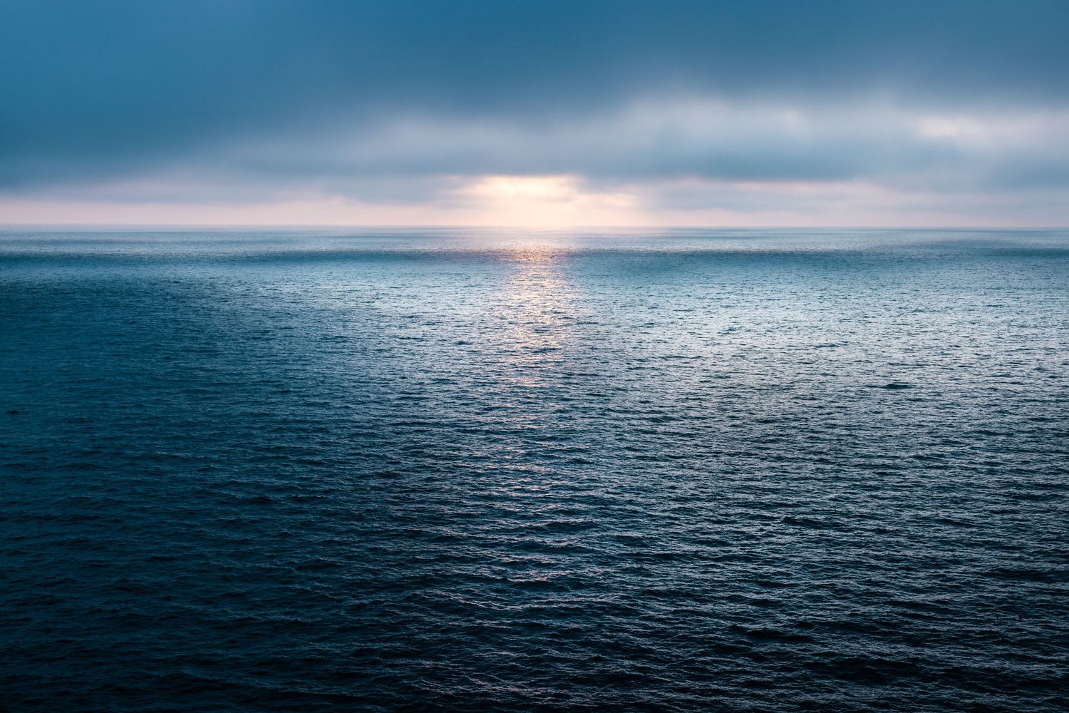 Tranquil ocean panorama at dusk with the sun setting over calm waters and cloudy horizon.