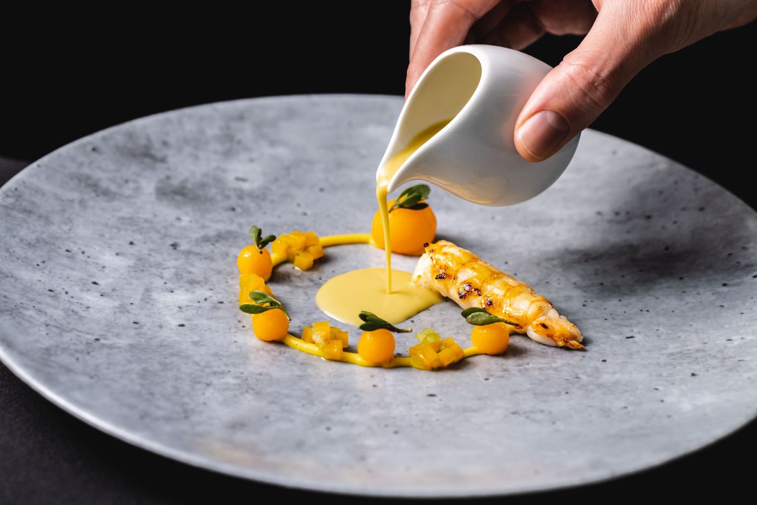 Hands drizzling sauce over artistically plated yellow garnishes on a gray plate.