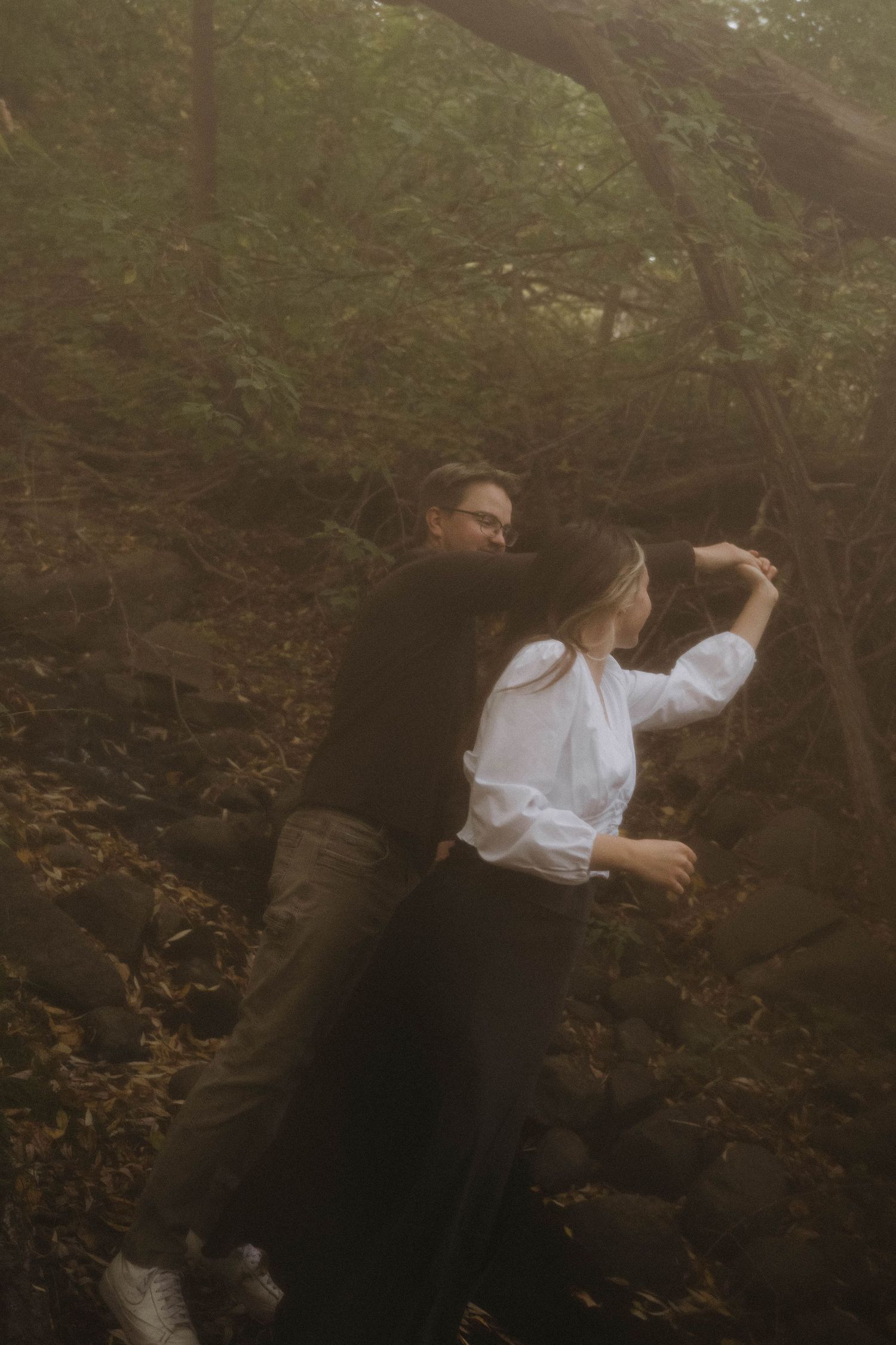 A couple dancing playfully among mossy rocks and lush green trees in a misty forest.