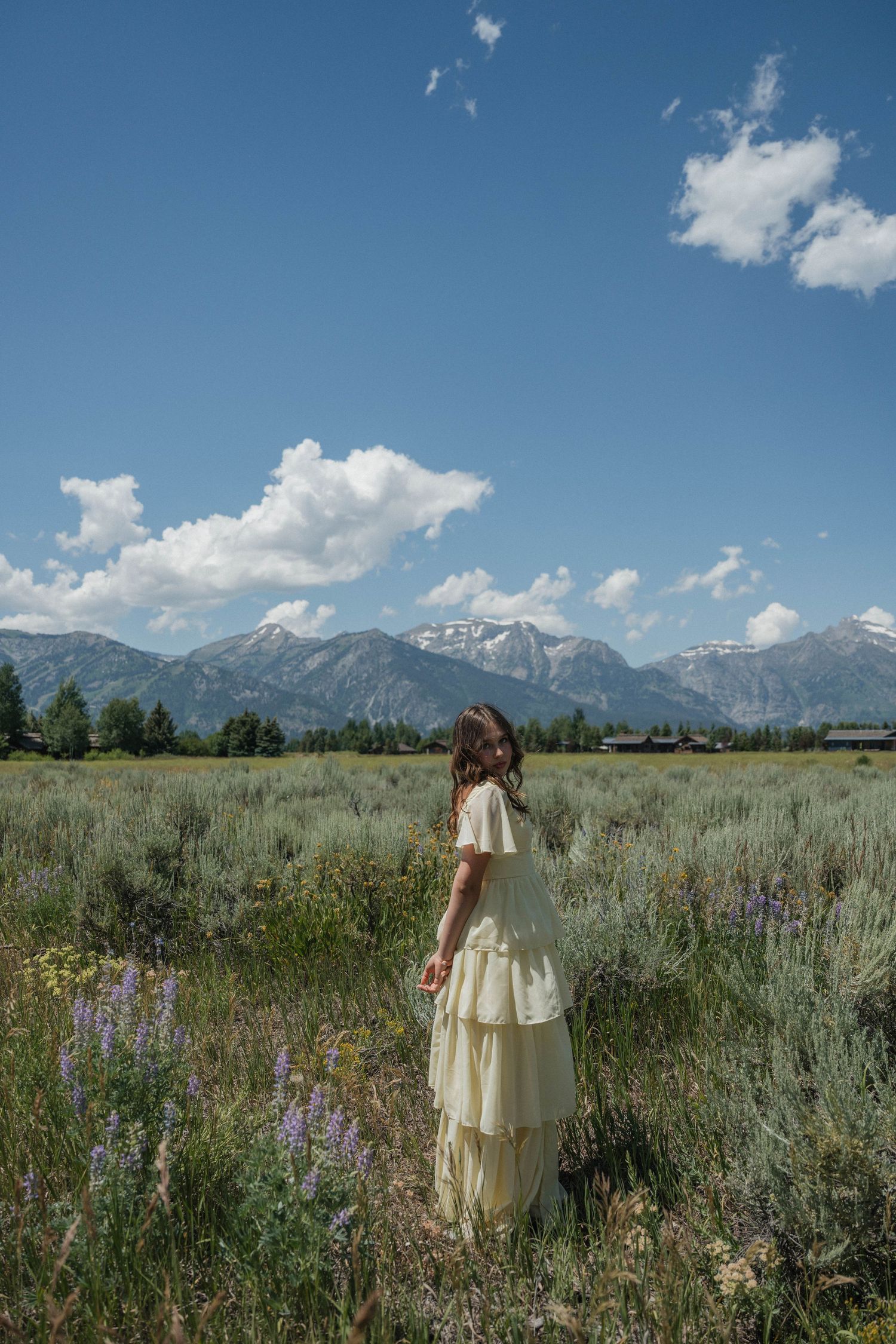Young woman in flowing yellow dress standing in wildflower meadow with snow-capped mountains behind.