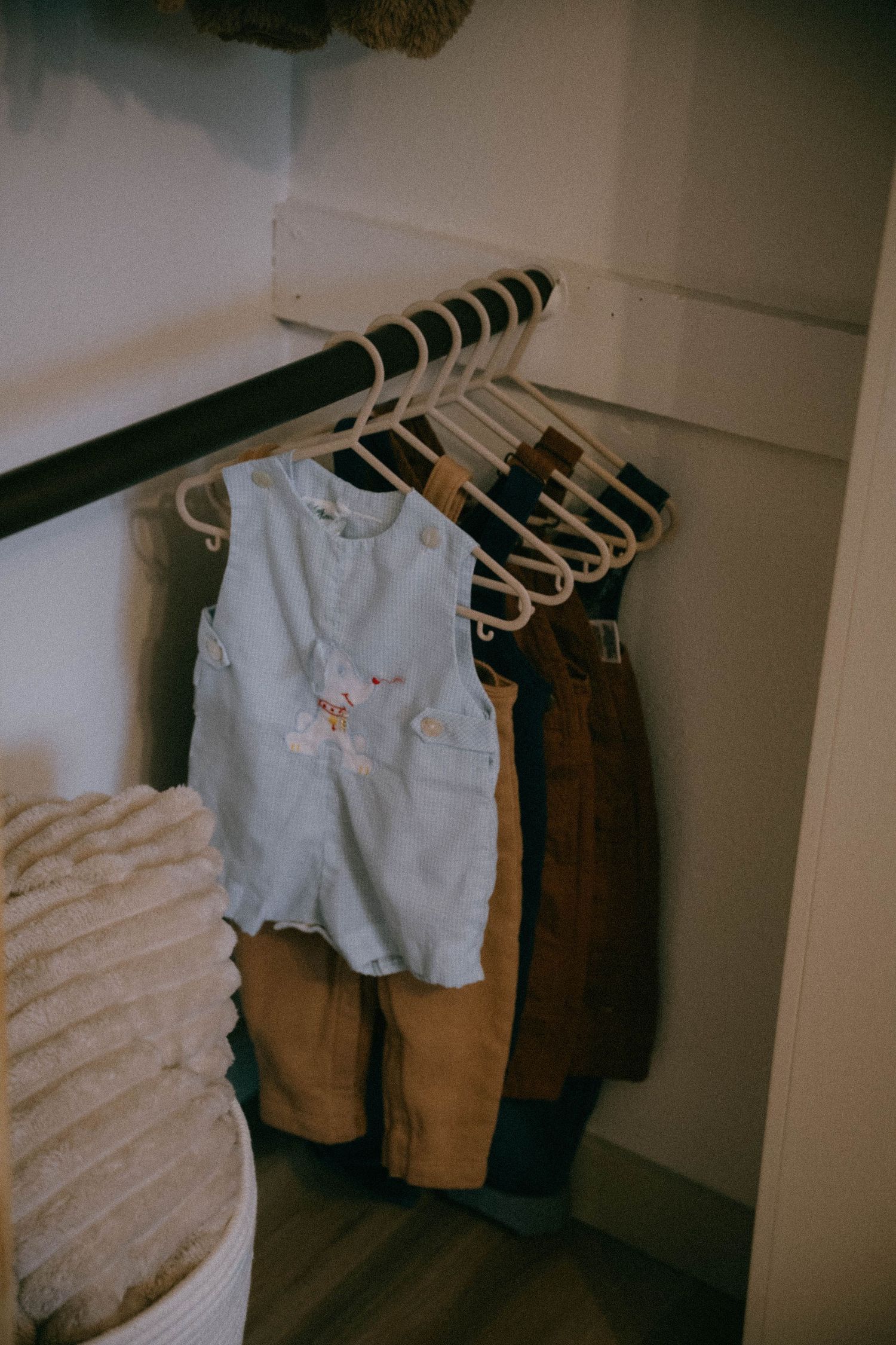 Small baby clothes including blue romper and brown pants hanging in nursery closet.