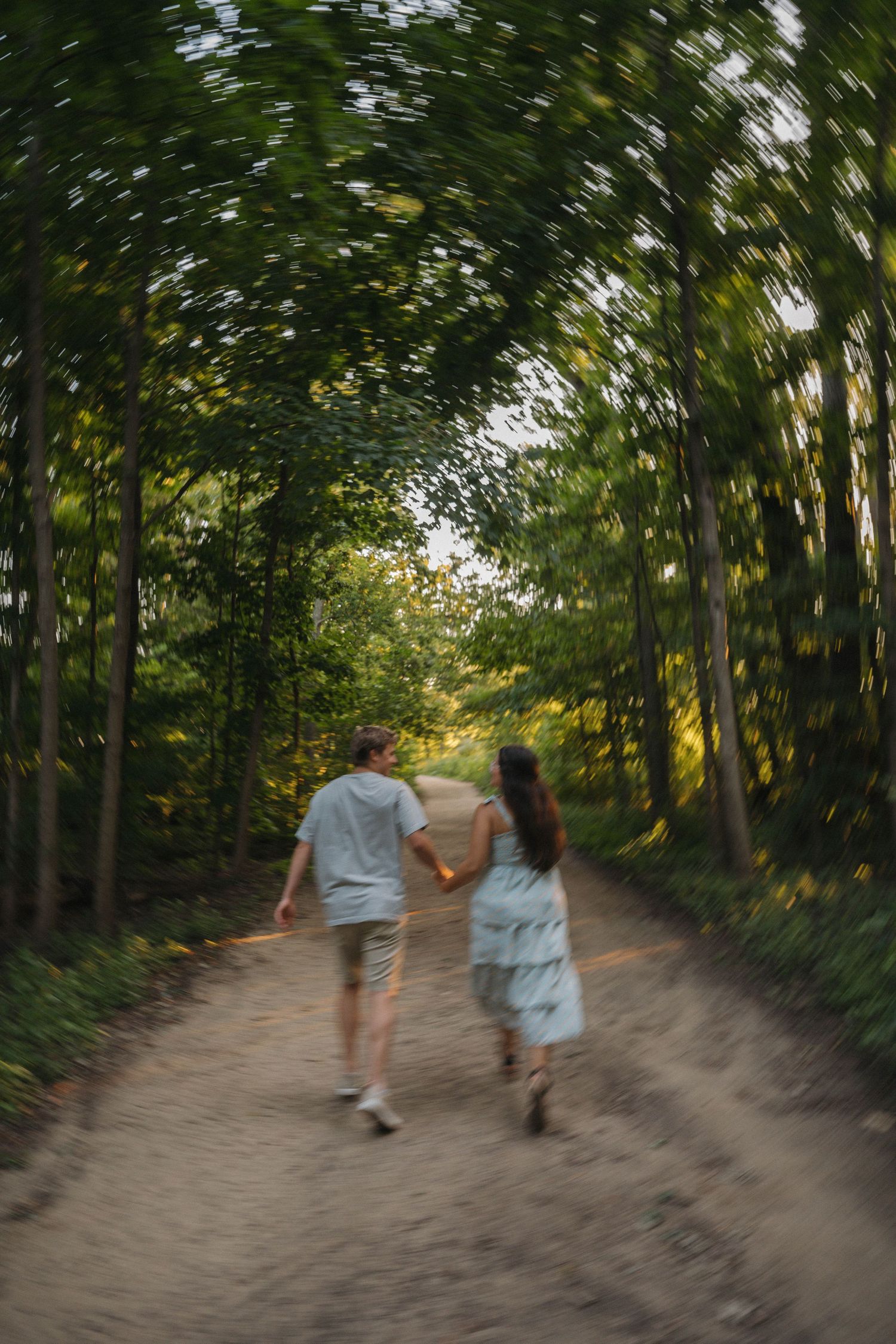 A couple holding hands while walking down a tree-lined dirt path in a lush green forest.