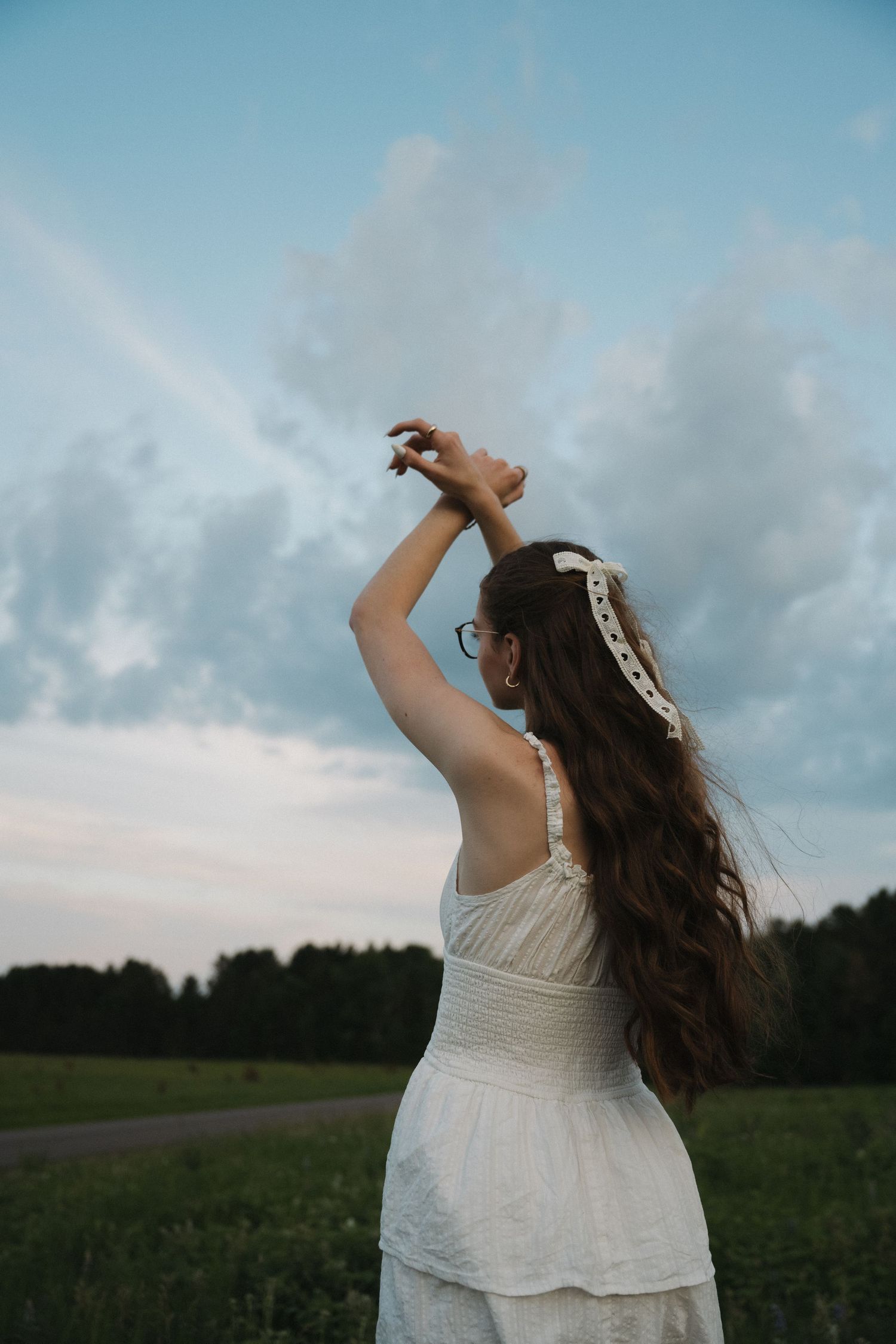 Young woman in white dress raises her arm gracefully in a green field under a dramatic cloudy sky at dusk.