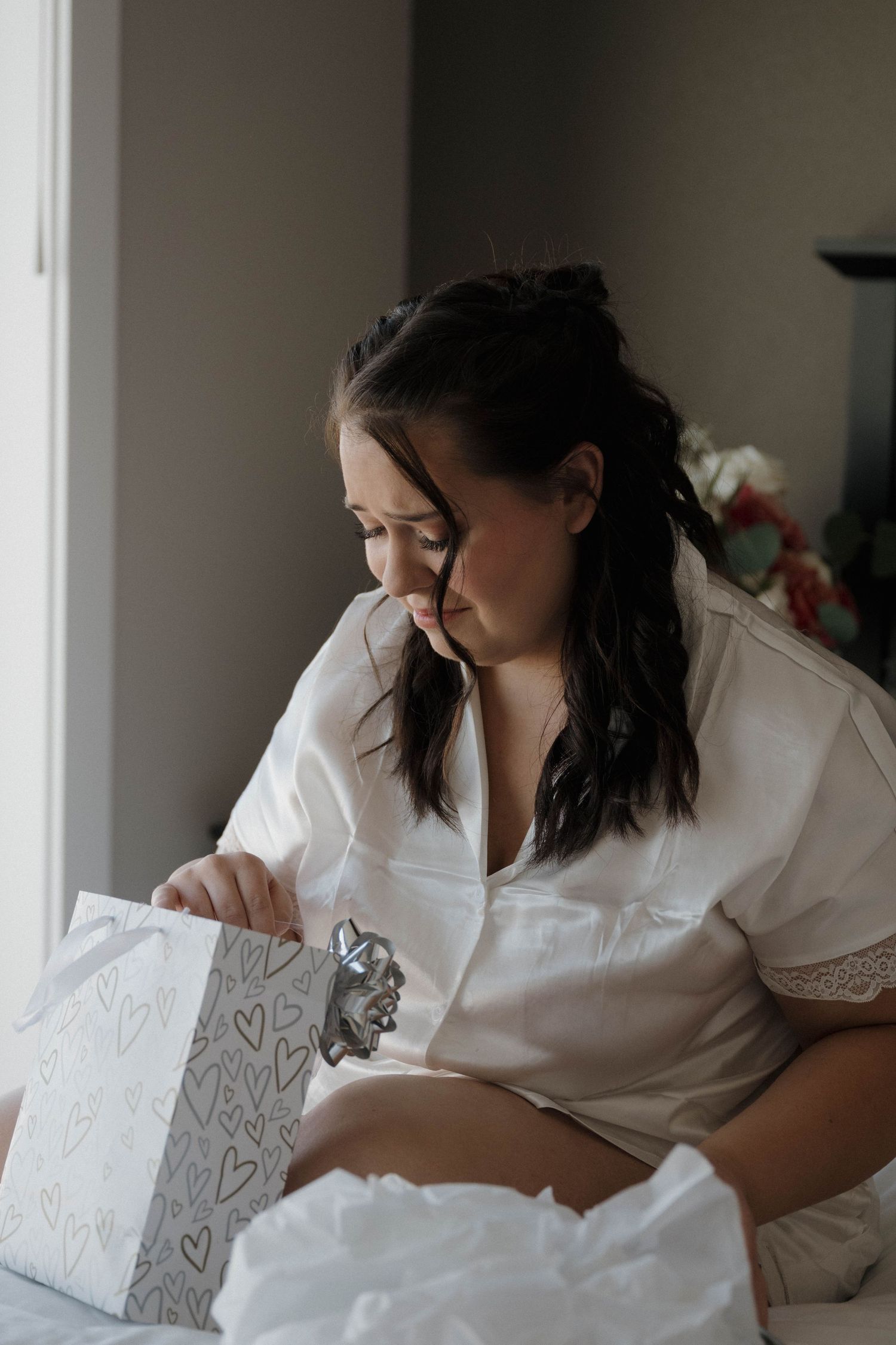 Bride in white satin robe sits on bed opening a gift bag with hearts beside a bright window.