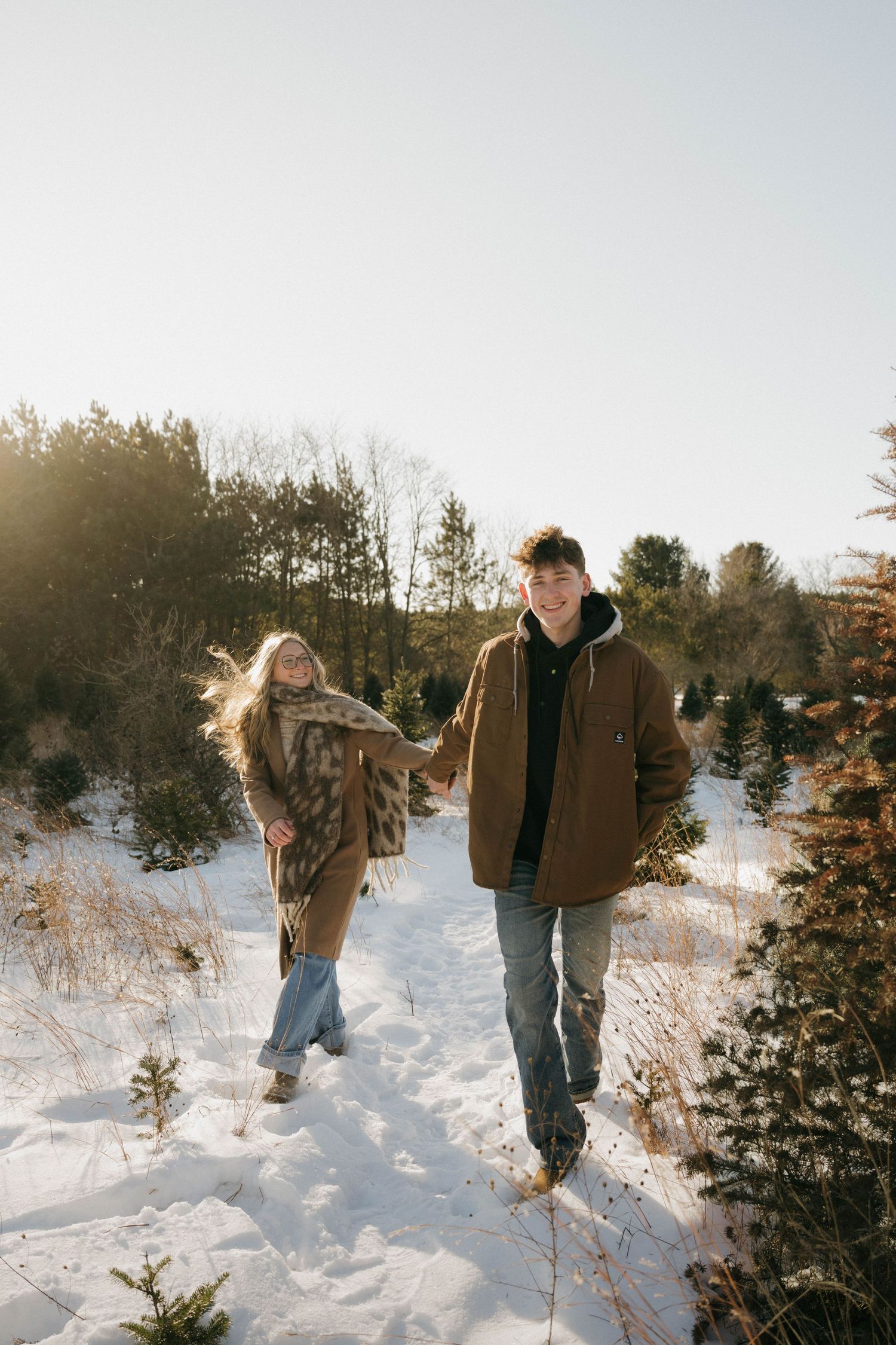 Couple holding hands walking through snowy Christmas tree farm path in bright winter sunlight.