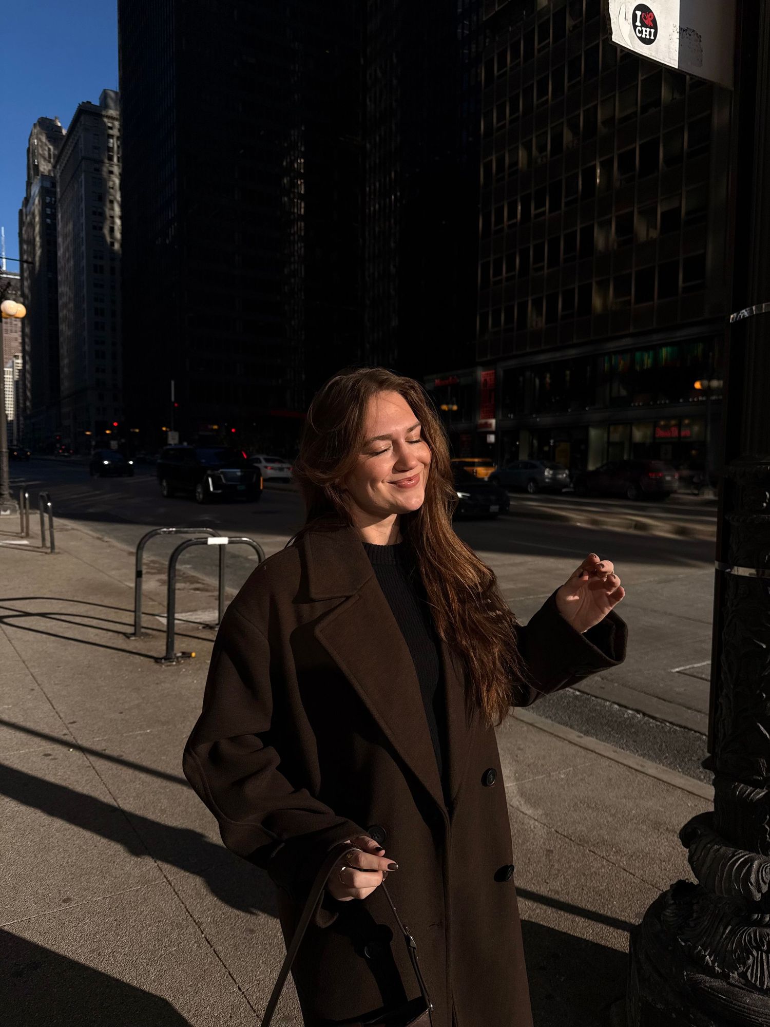 Young woman in brown coat smiles in golden sunlight on a busy city sidewalk.