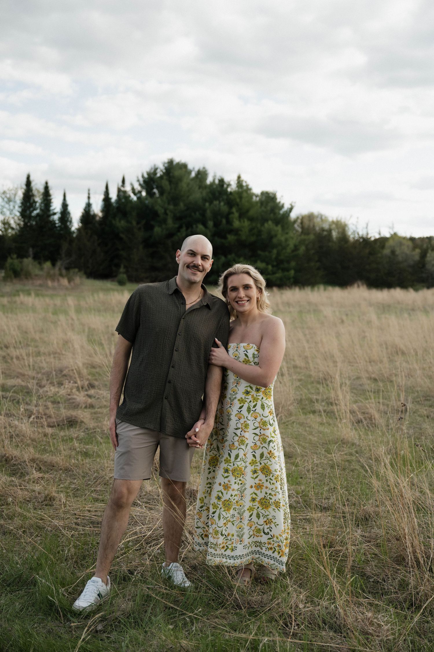 Couple smiling and embracing in a golden field with evergreen trees, woman in floral dress, man in olive shirt.