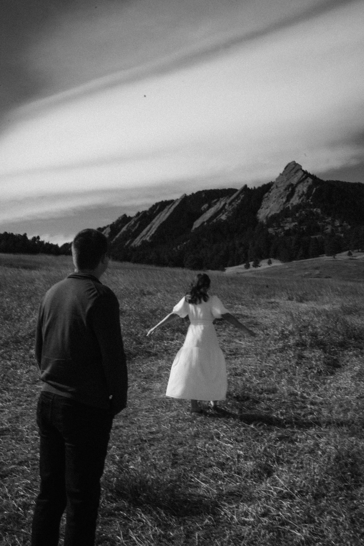 Black and white photo of couple walking toward dramatic Flatirons rock formations in Boulder Colorado field.