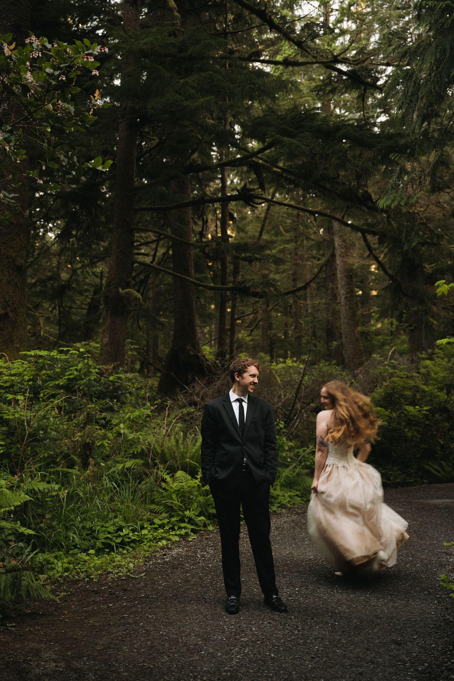 Groom in black suit watches bride in flowing dress twirl on forest path surrounded by tall trees.