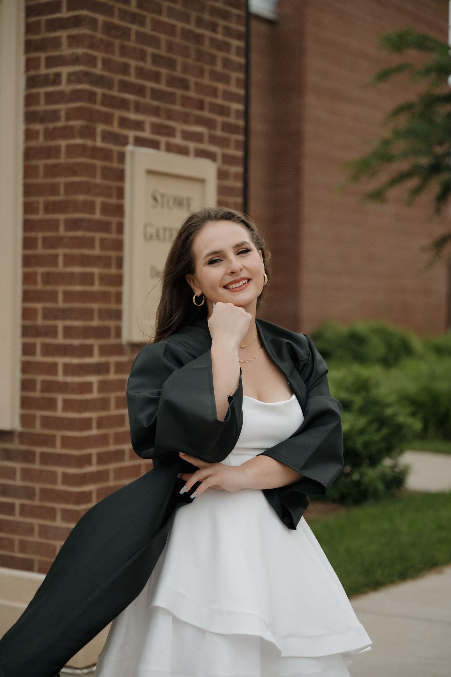Young woman in white dress and black jacket smiling outdoors near brick building at Stowe Gate.
