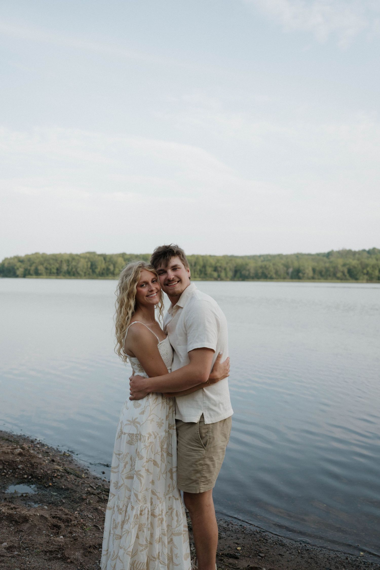 A smiling couple embraces on a lakeside shore, woman in white floral dress, man in casual shirt and khaki shorts.