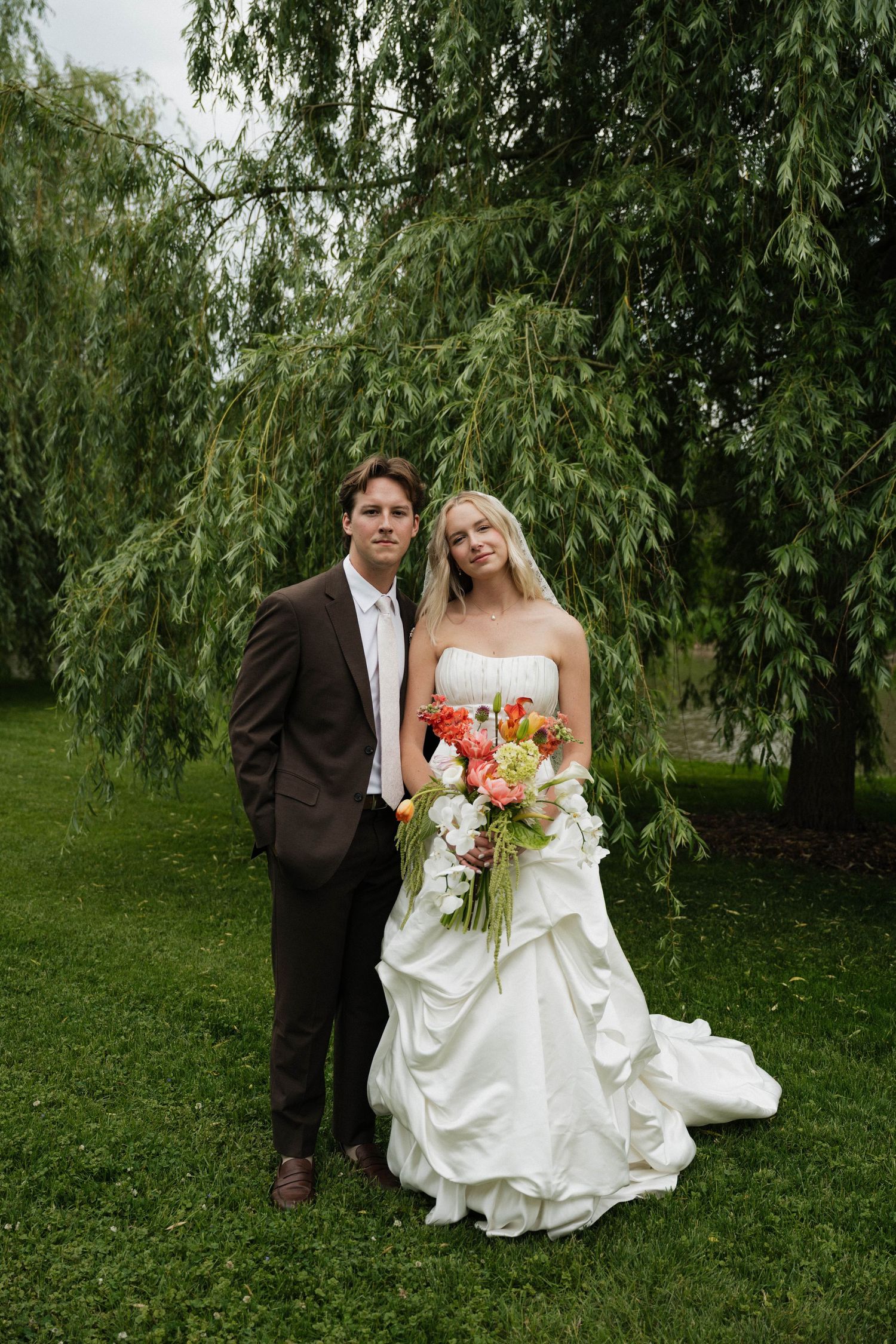 Bride in strapless white gown holding colorful bouquet stands with groom in brown suit under weeping willow tree.