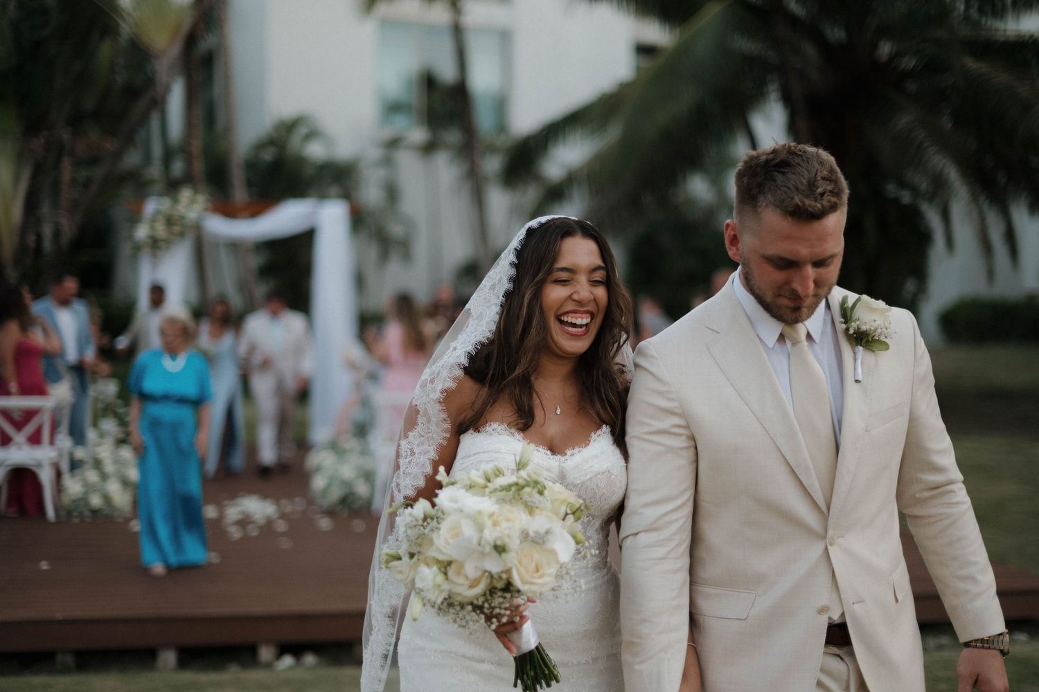 Radiant bride laughing with groom in beige suit after outdoor tropical wedding ceremony with white florals.