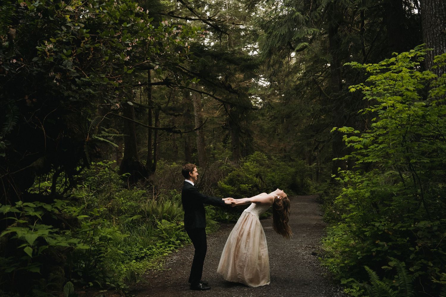 Groom dips bride dramatically on a forest path surrounded by tall green trees in a moody woodland setting.